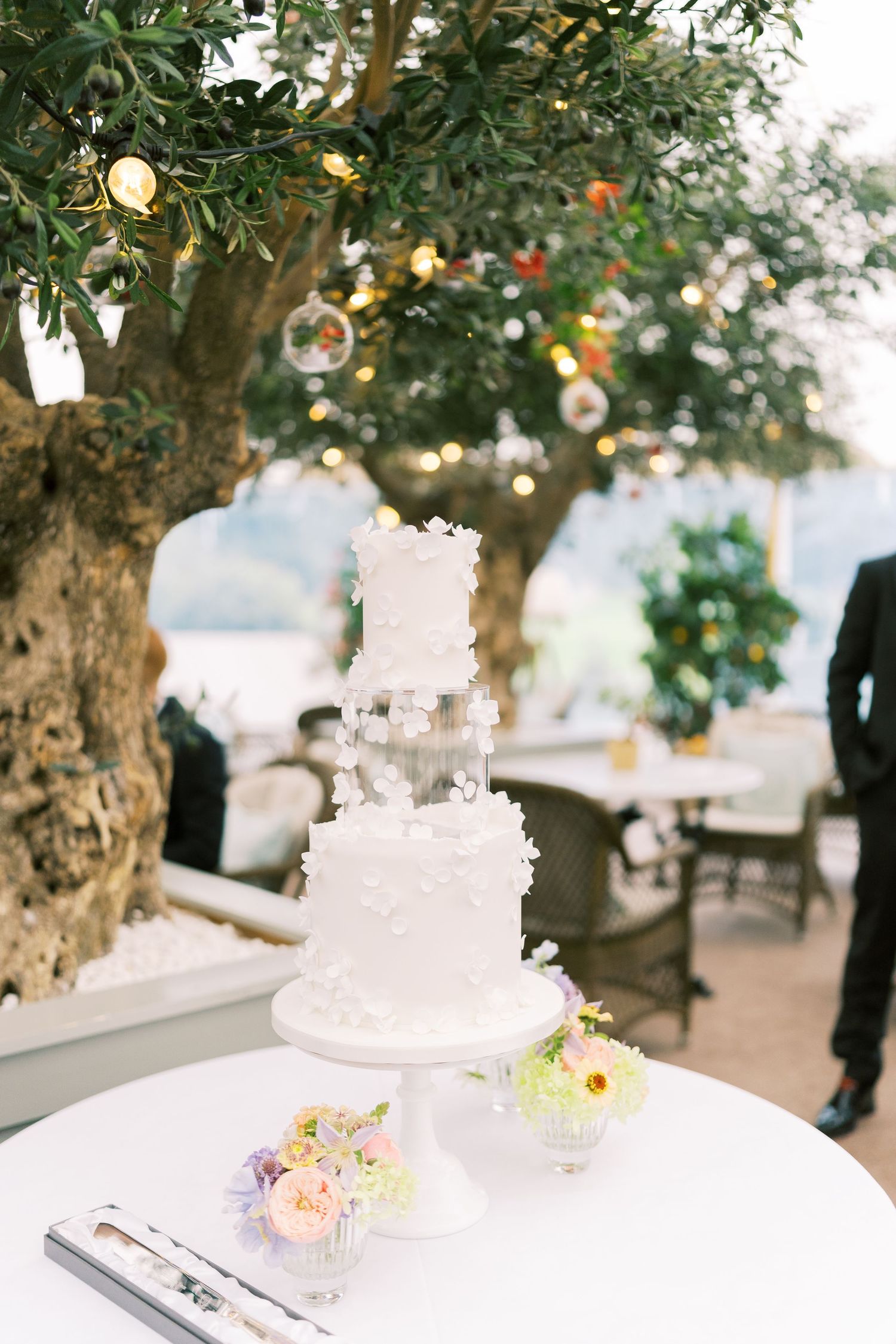 Elegant three-tier white wedding cake displayed on table beneath olive tree with fairy lights outdoors.