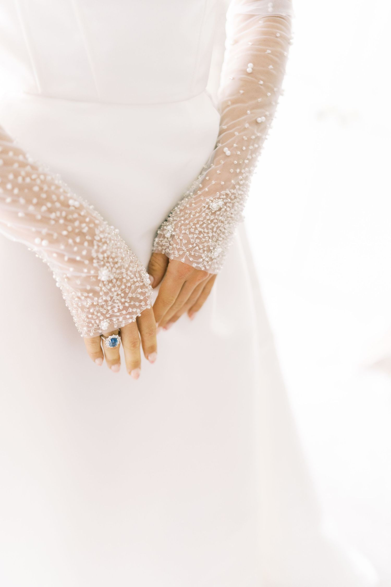 Close-up of bride's hands adorned with pearl-embellished sheer gloves and a sapphire ring against white wedding dress.