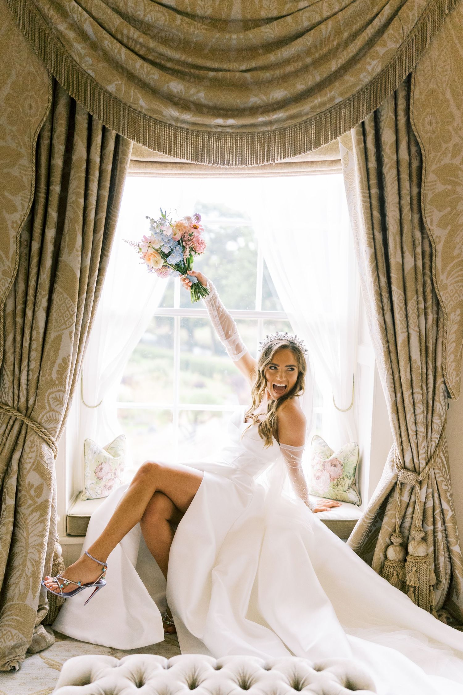 Joyful bride in a white wedding dress and tiara raising her bouquet while seated in an elegant window alcove.