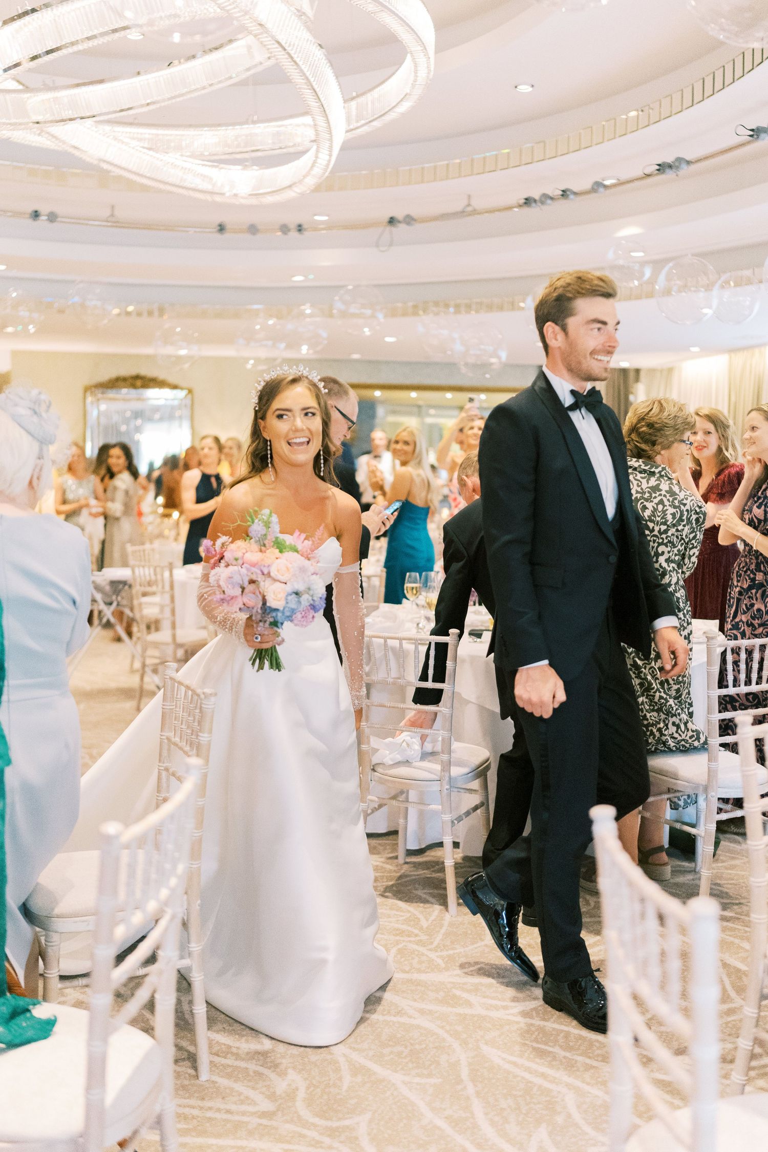 Bride and groom walk down the aisle at elegant indoor wedding reception, bride holding pink and blue floral bouquet.