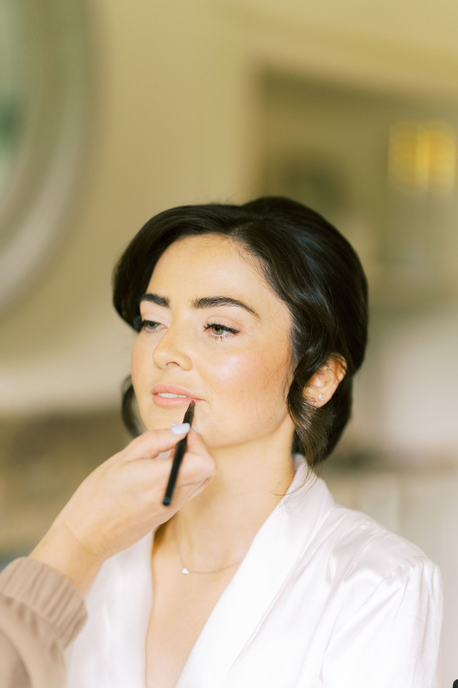 Bride getting lip makeup applied with a brush while wearing a white robe during wedding preparations.