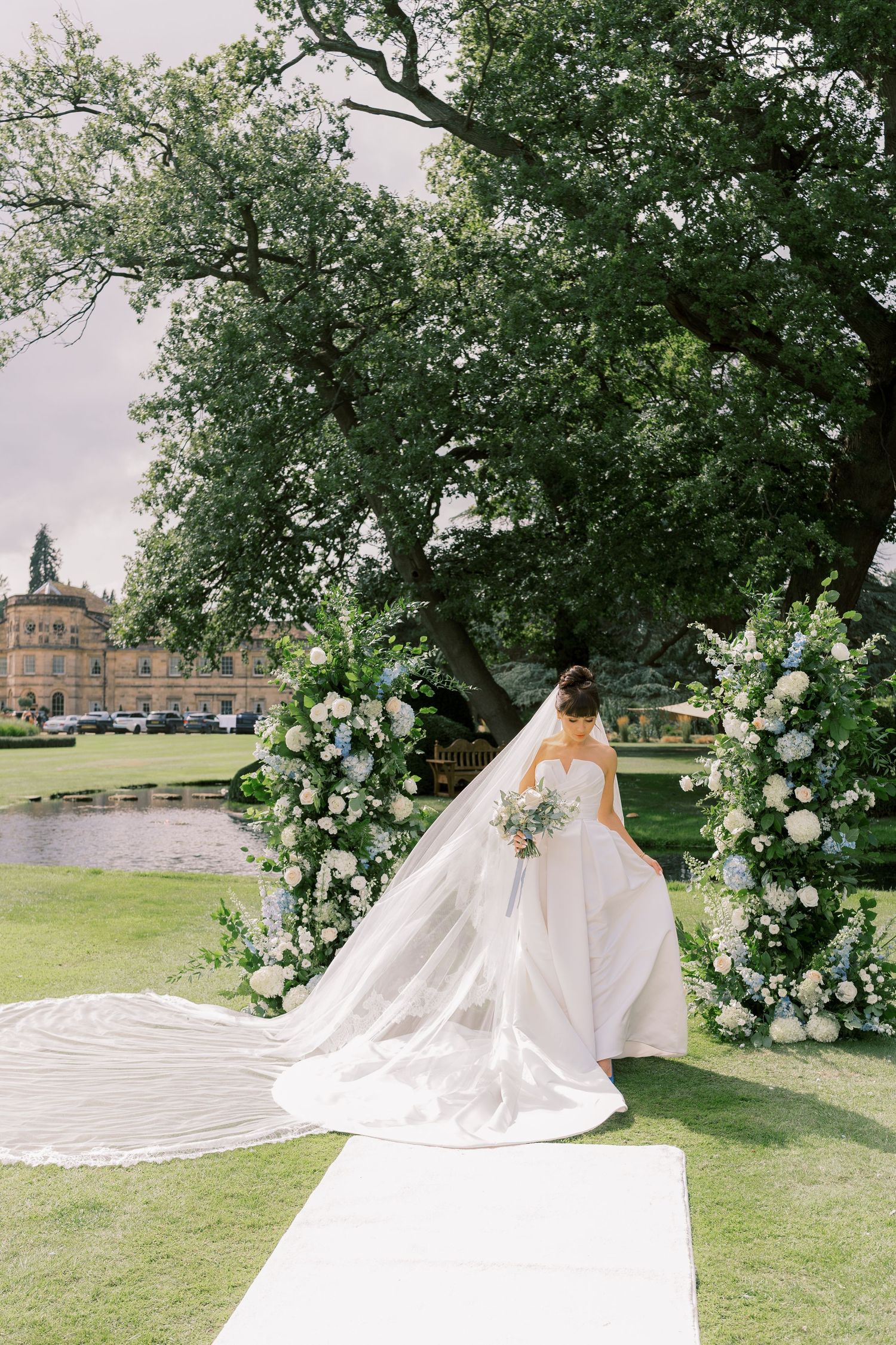 Bride in a flowing white gown with long veil stands at floral arch altar outdoors near a grand estate.