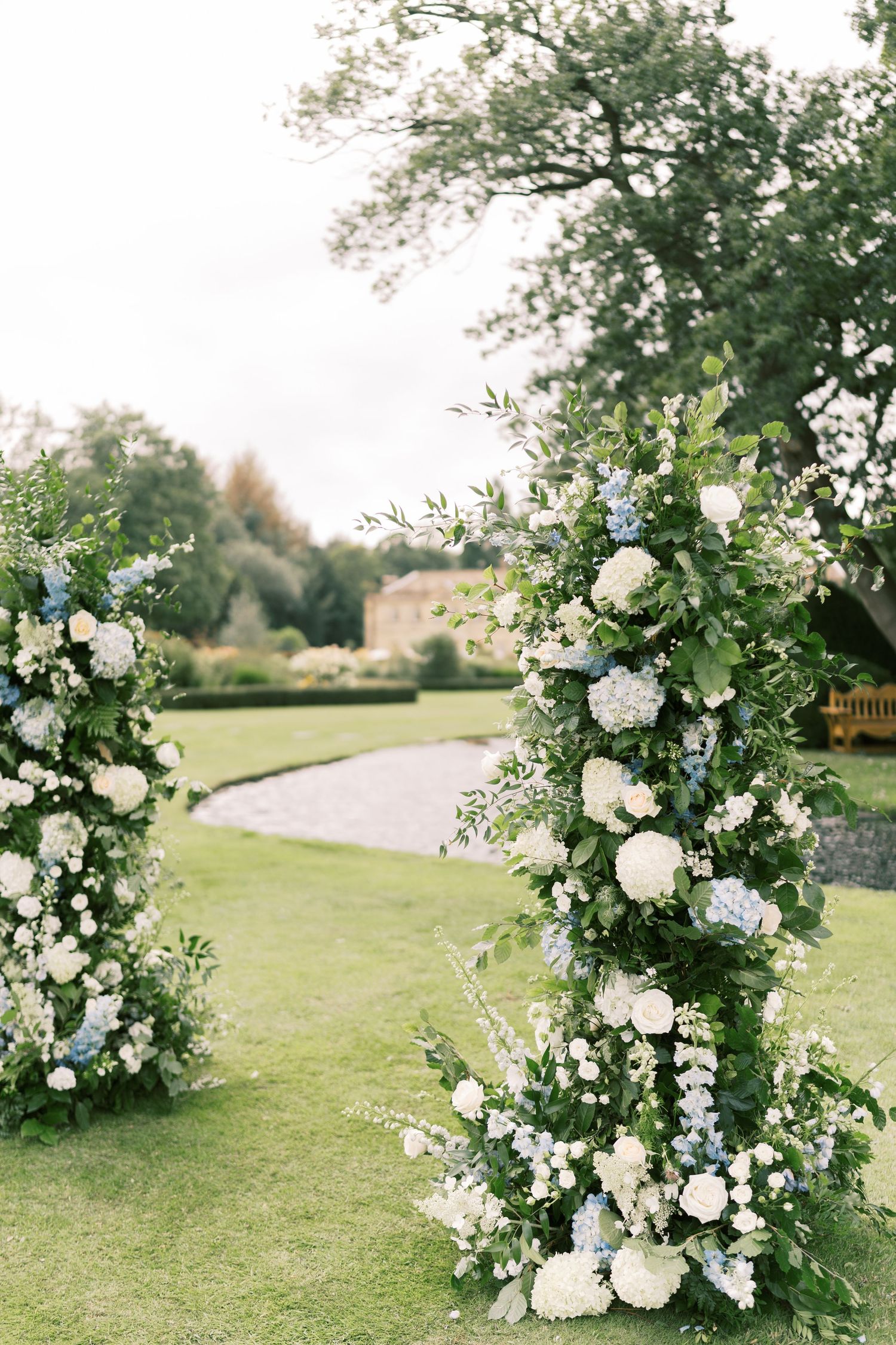 Tall floral arch columns with white hydrangeas and blue flowers frame an outdoor garden wedding ceremony aisle.