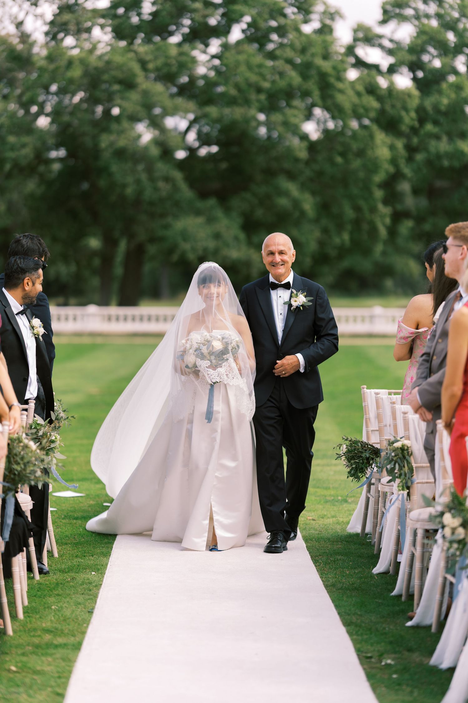 Bride in white gown and veil walks down outdoor aisle with escort at garden wedding ceremony.