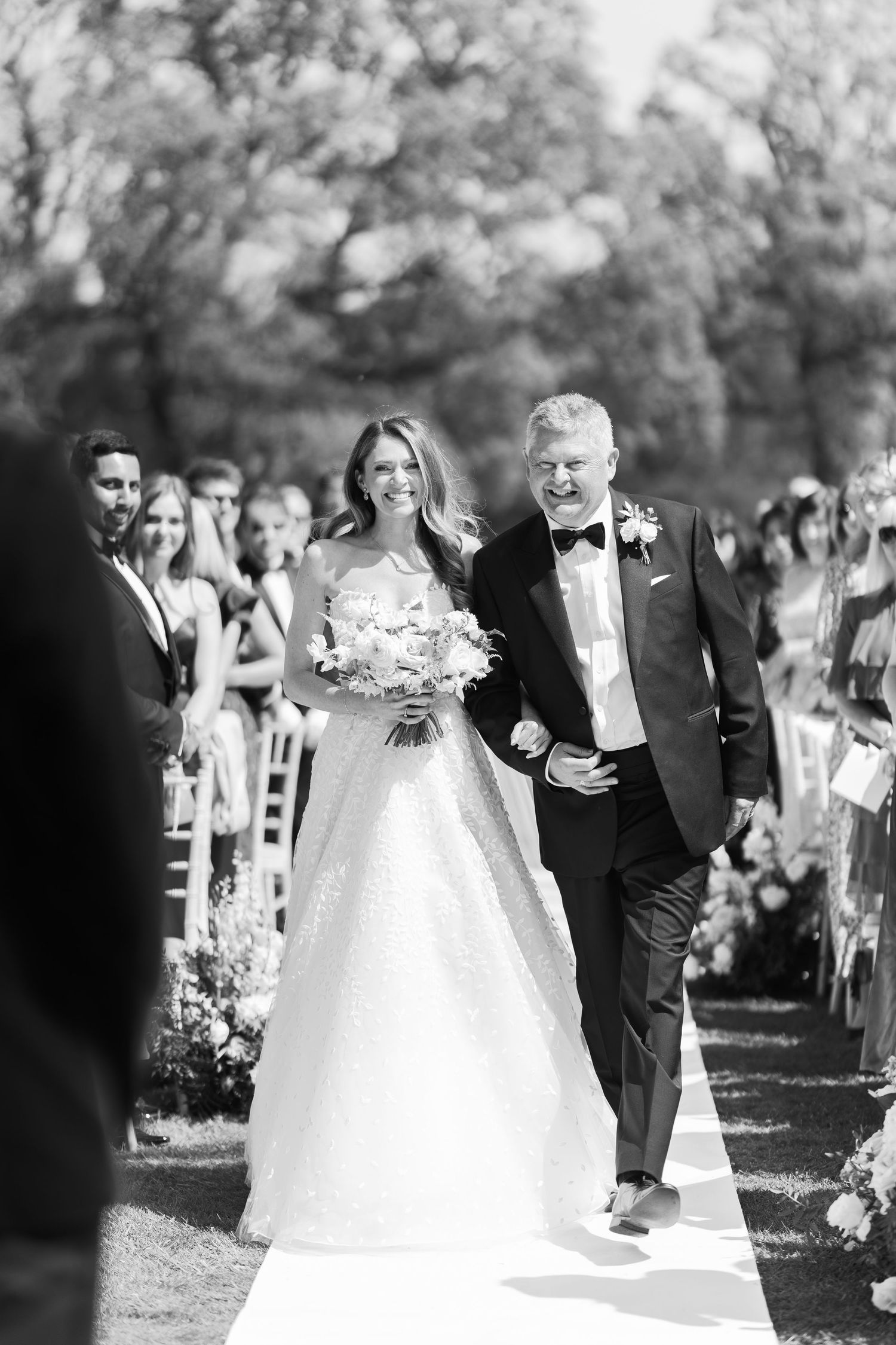 Bride in sparkling ball gown walks down aisle with father in black tuxedo at elegant outdoor wedding.