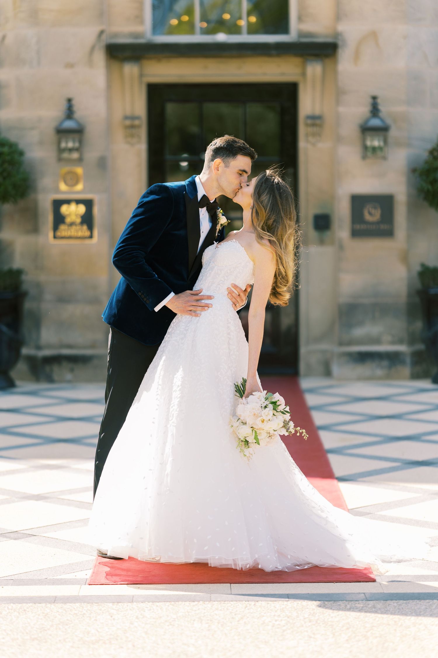 Bride and groom share a romantic kiss on a red carpet outside an elegant venue, bride holding a white bouquet.