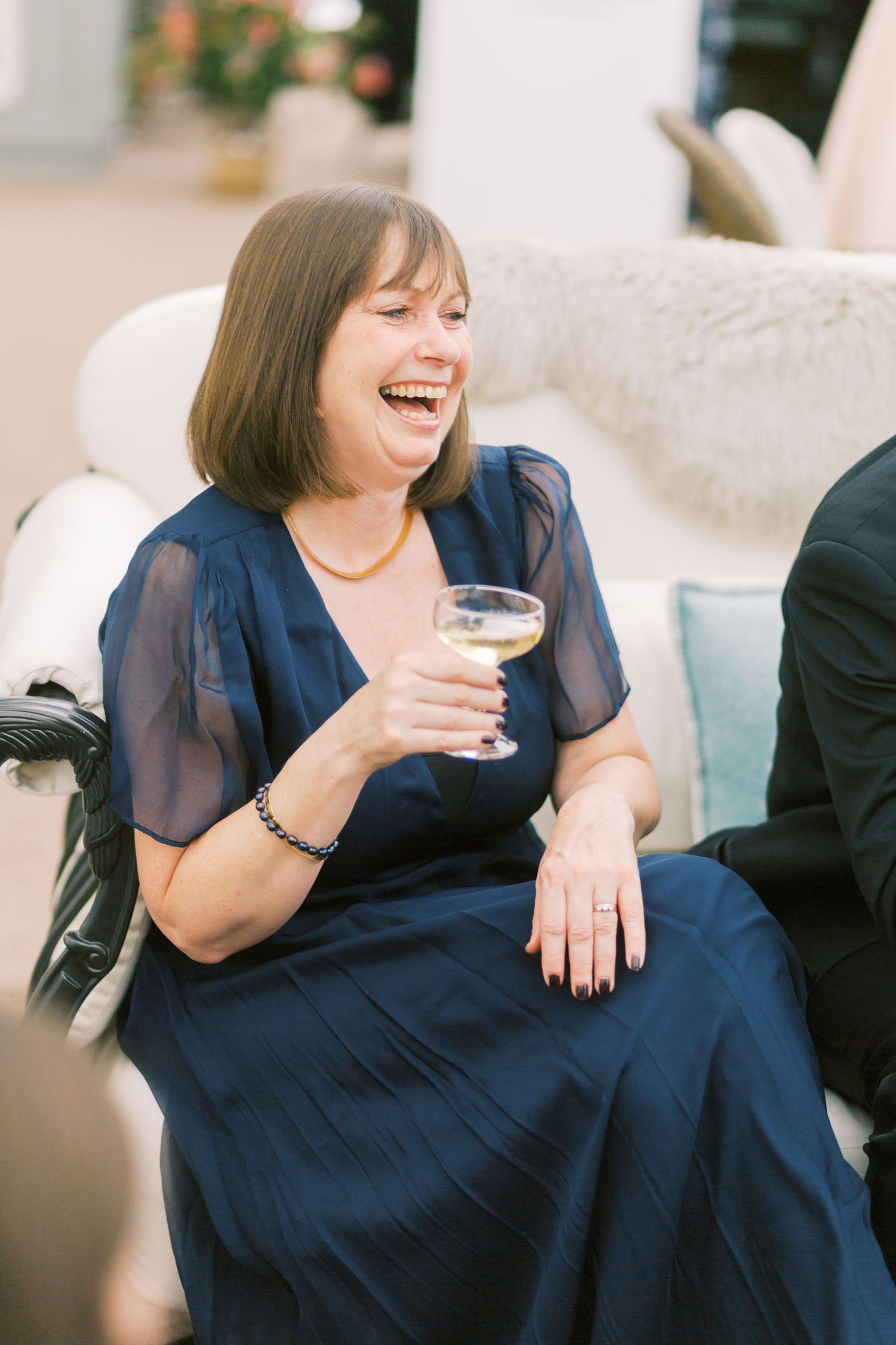A woman in a navy blue dress laughs joyfully while holding a glass of white wine at an elegant event.