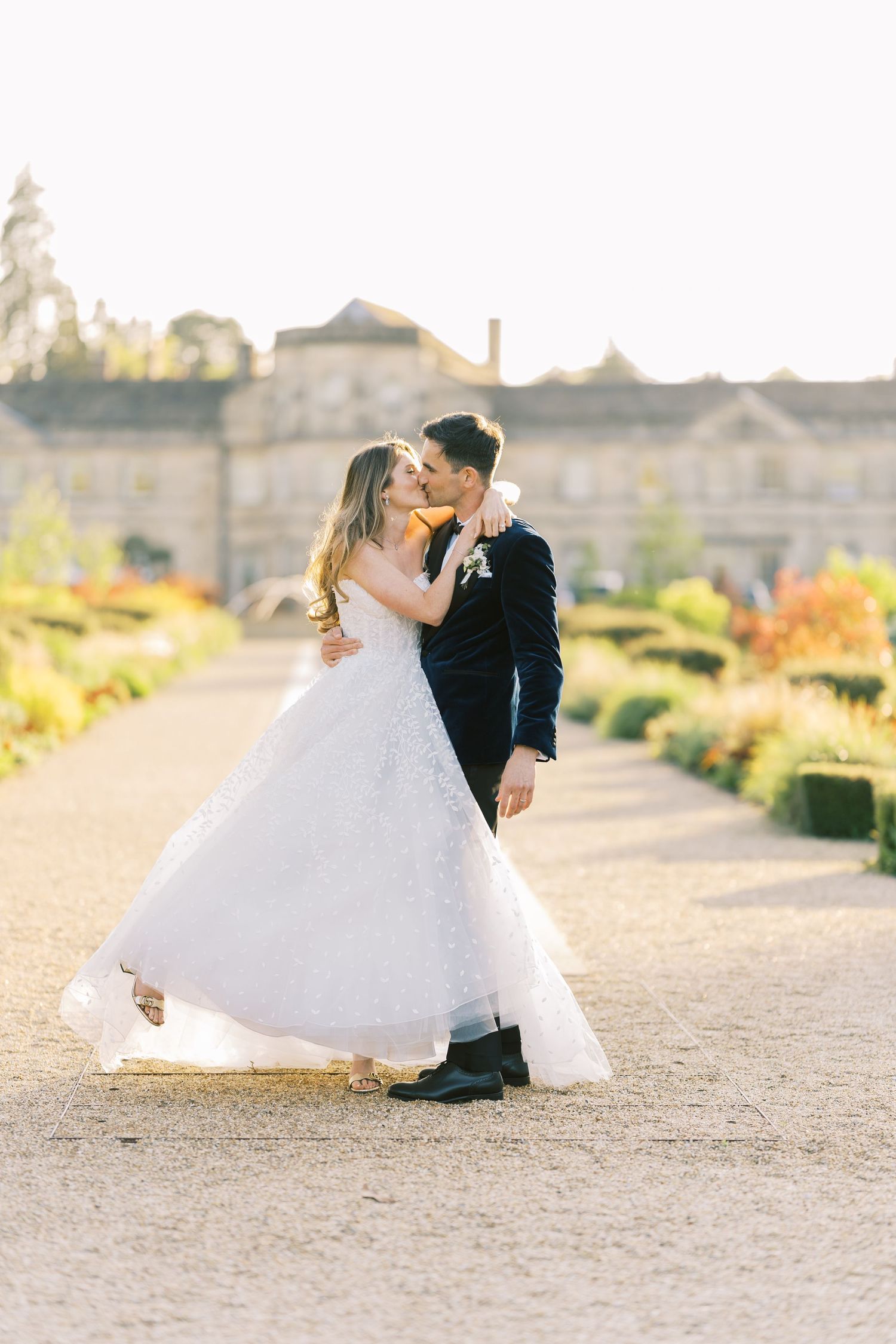 Bride and groom share a romantic kiss in a sunlit garden pathway with manicured hedges and a grand estate in the background.