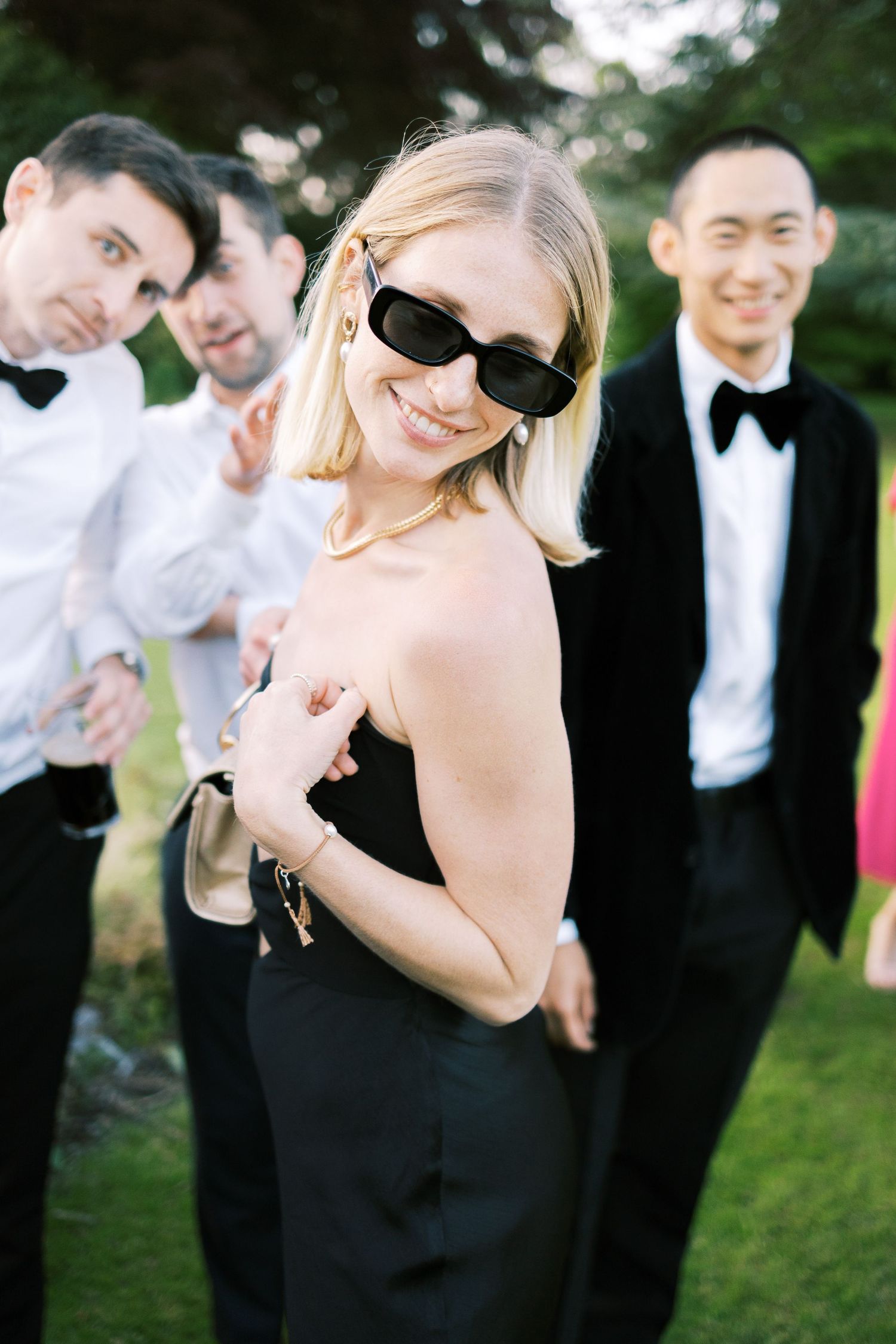 Blonde woman in black strapless dress and sunglasses smiles outdoors with men in tuxedos at a formal event.