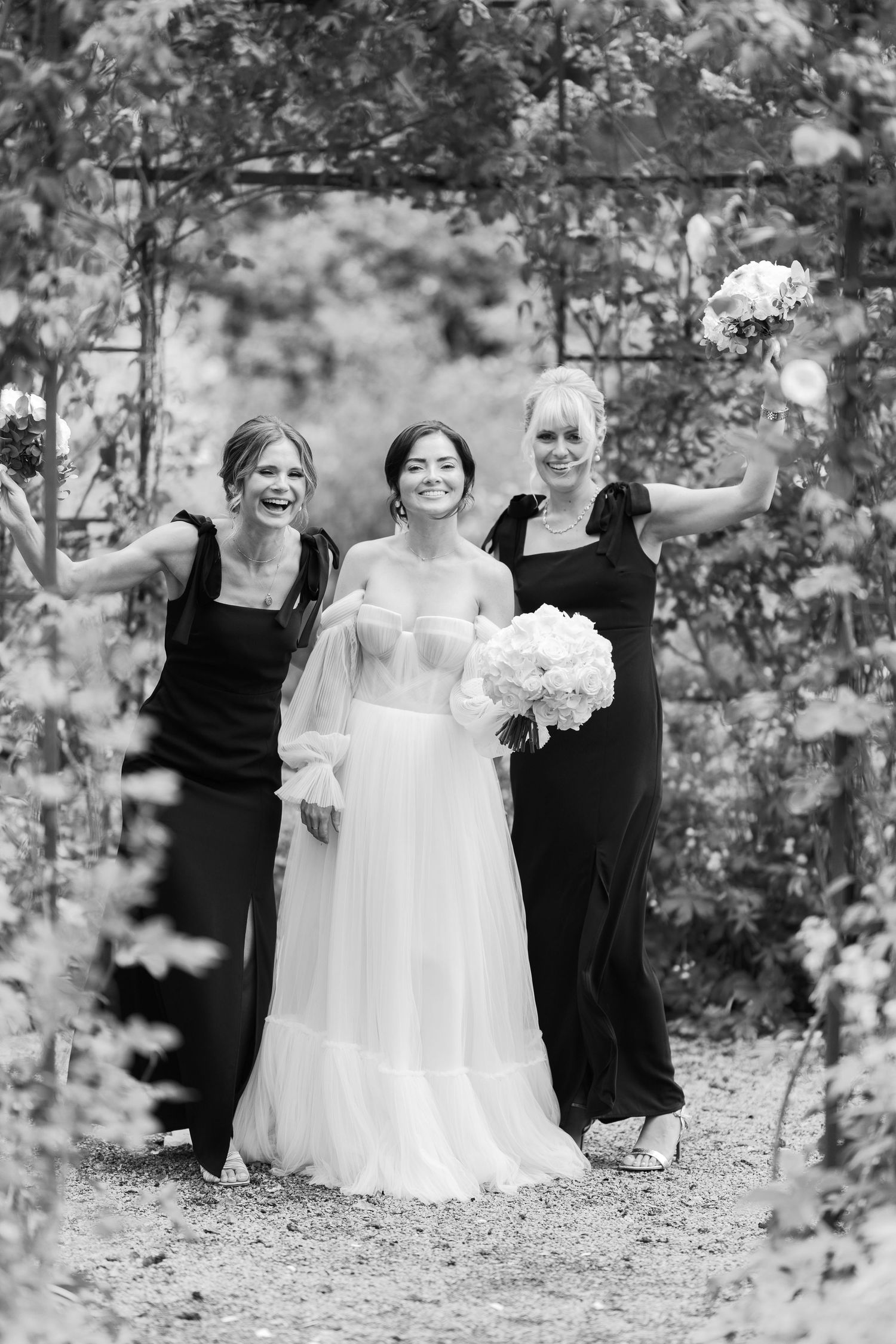 Bride in white gown poses with two bridesmaids in black dresses under a floral arch in black and white photo.