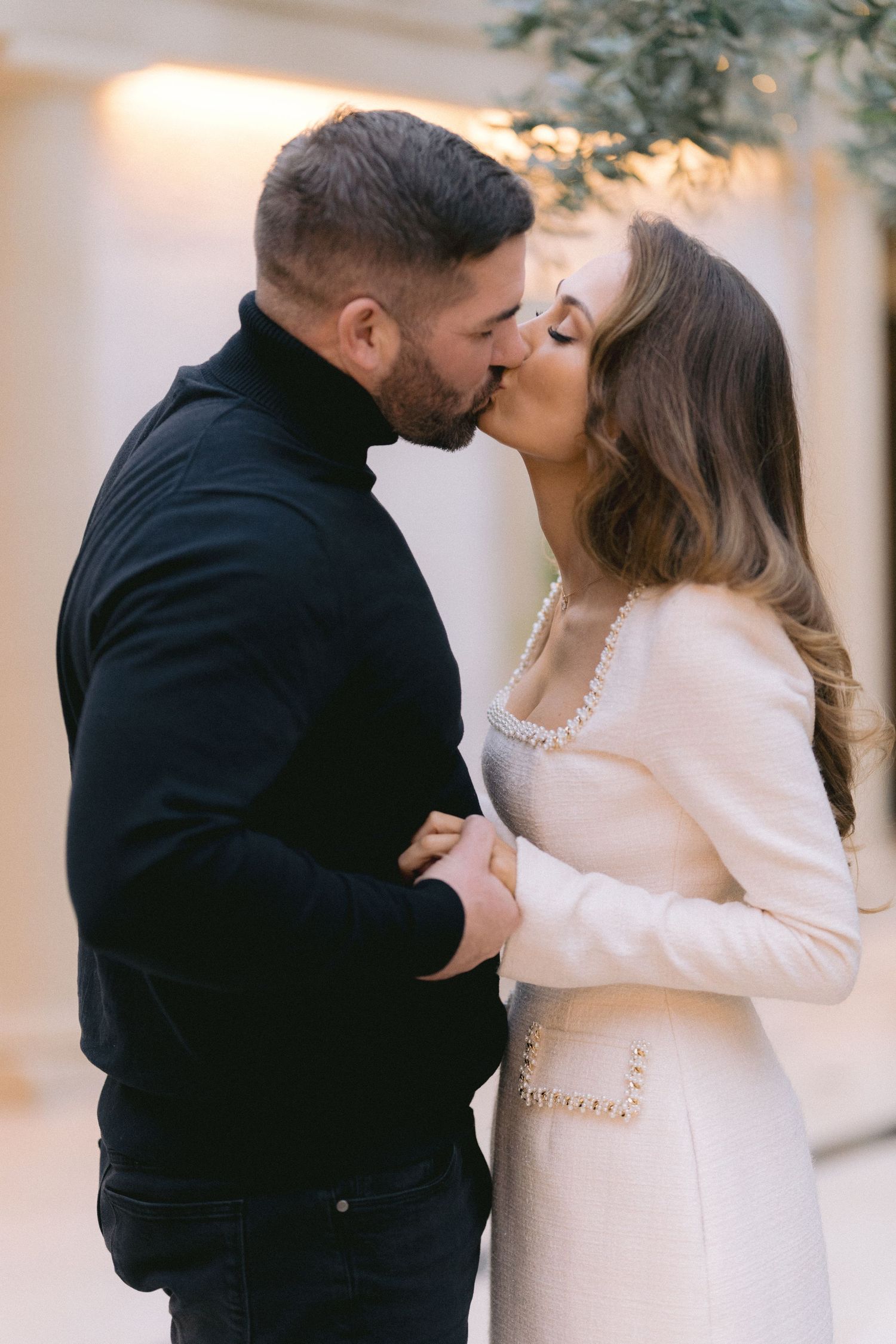 Engaged couple sharing a romantic kiss, man in black turtleneck and woman in elegant white coat.