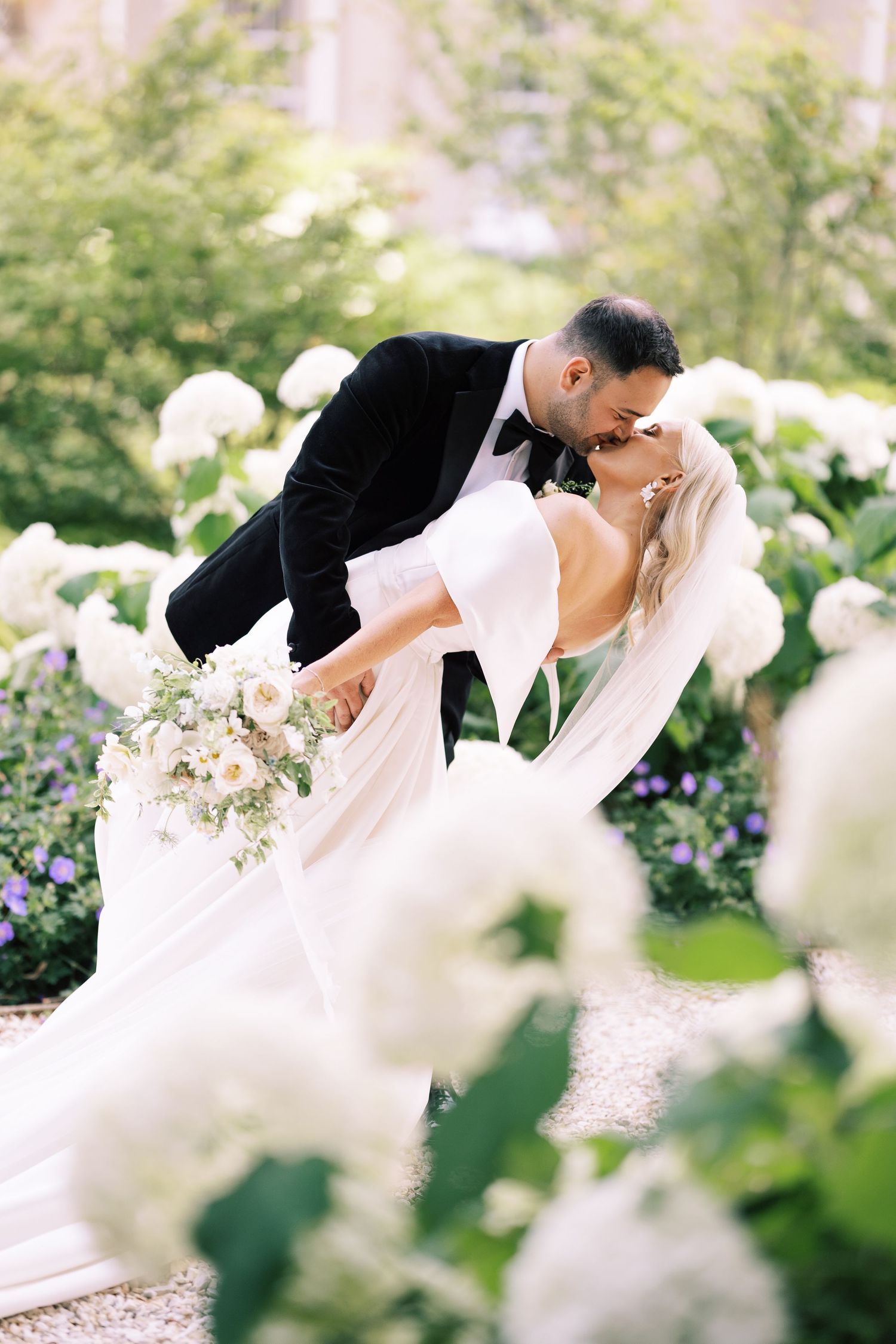 Couple sharing a romantic dip kiss framed by blooming white hydrangea bushes.