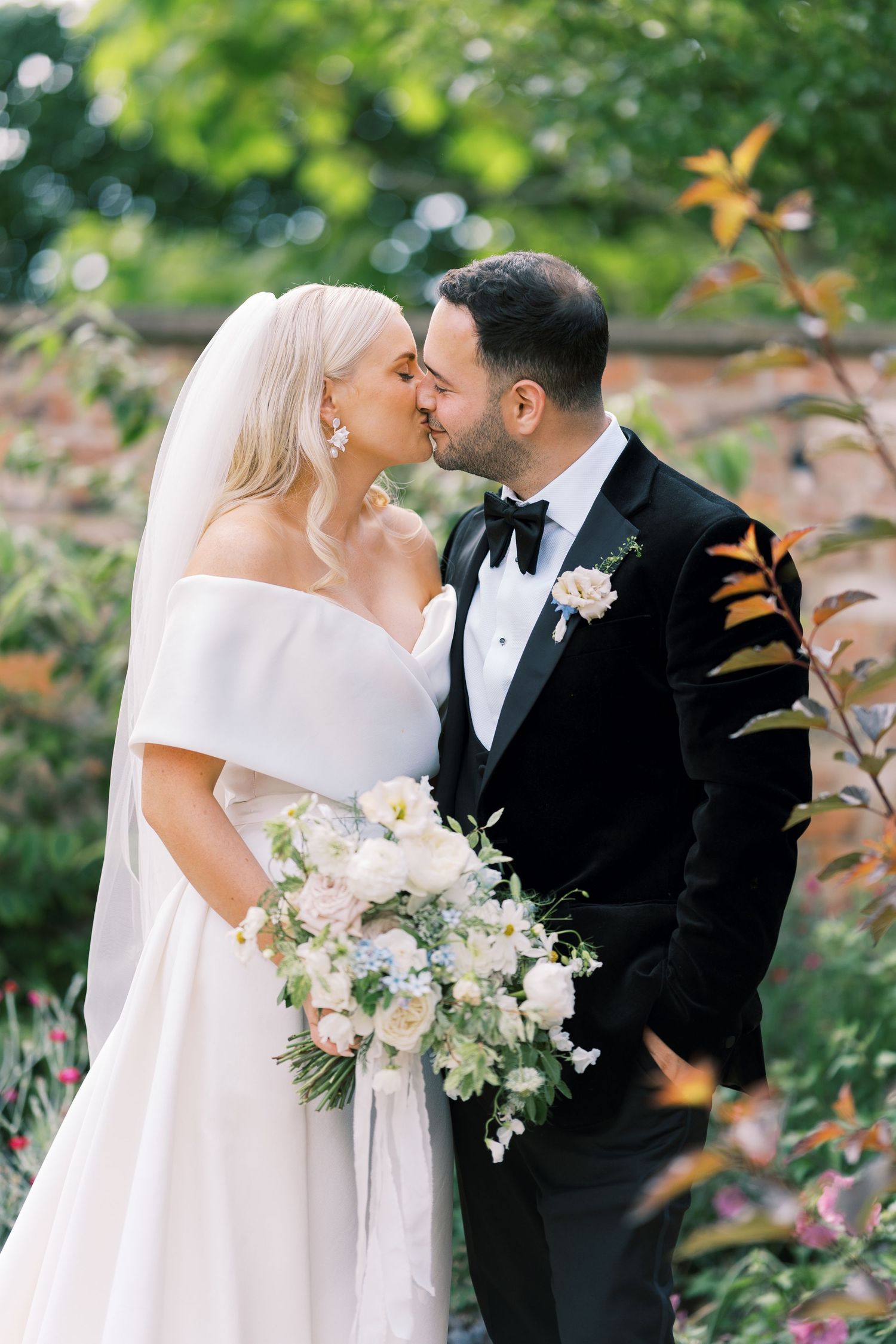 Bride and groom sharing a kiss in a lush garden, bride holding elegant white floral bouquet.