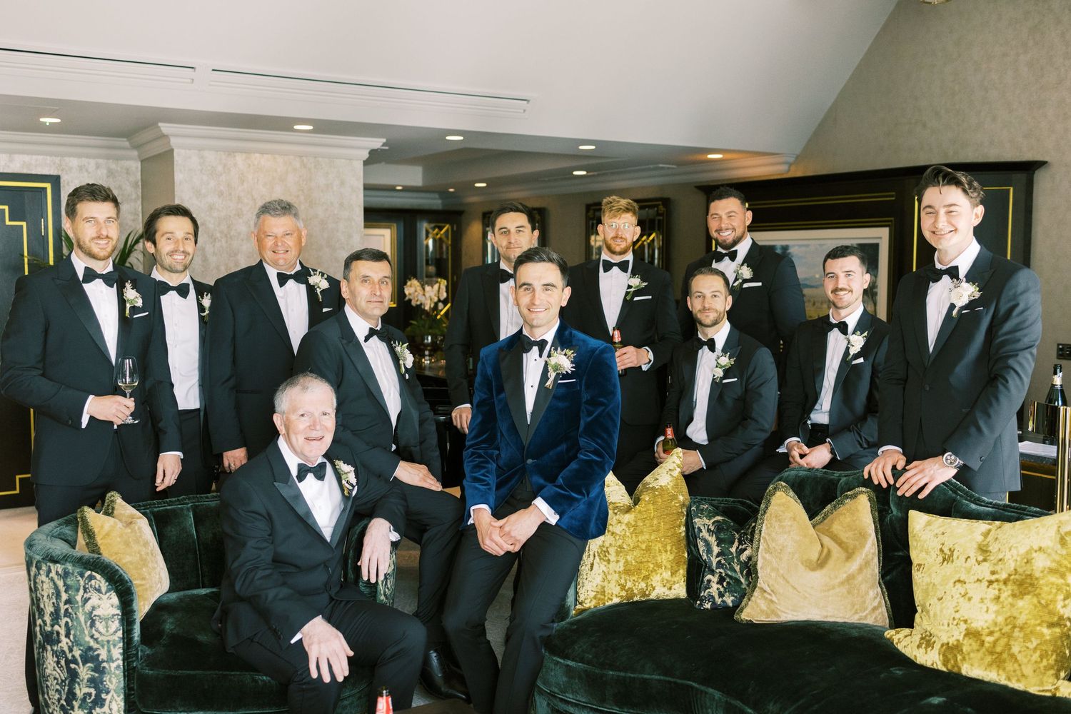 Groom in blue velvet tuxedo poses with groomsmen in black tuxedos in an elegant lounge setting.