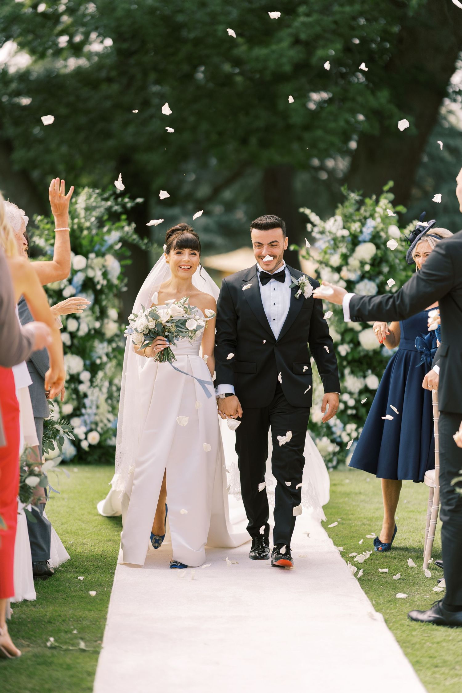 Bride and groom walk down the aisle as guests toss white flower petals at outdoor garden wedding ceremony.