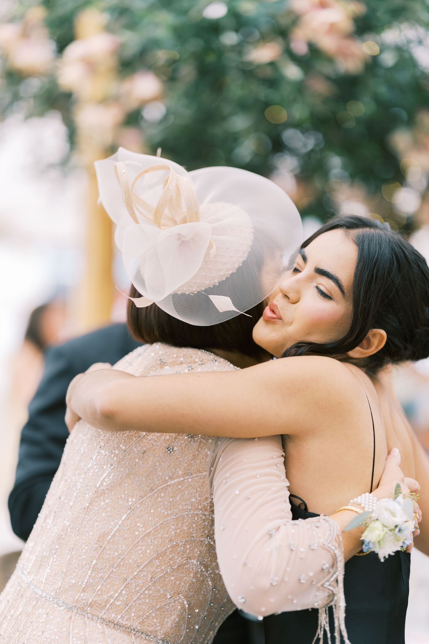 Two women share a warm embrace at a wedding, one wearing a elegant fascinator hat and sparkly dress.