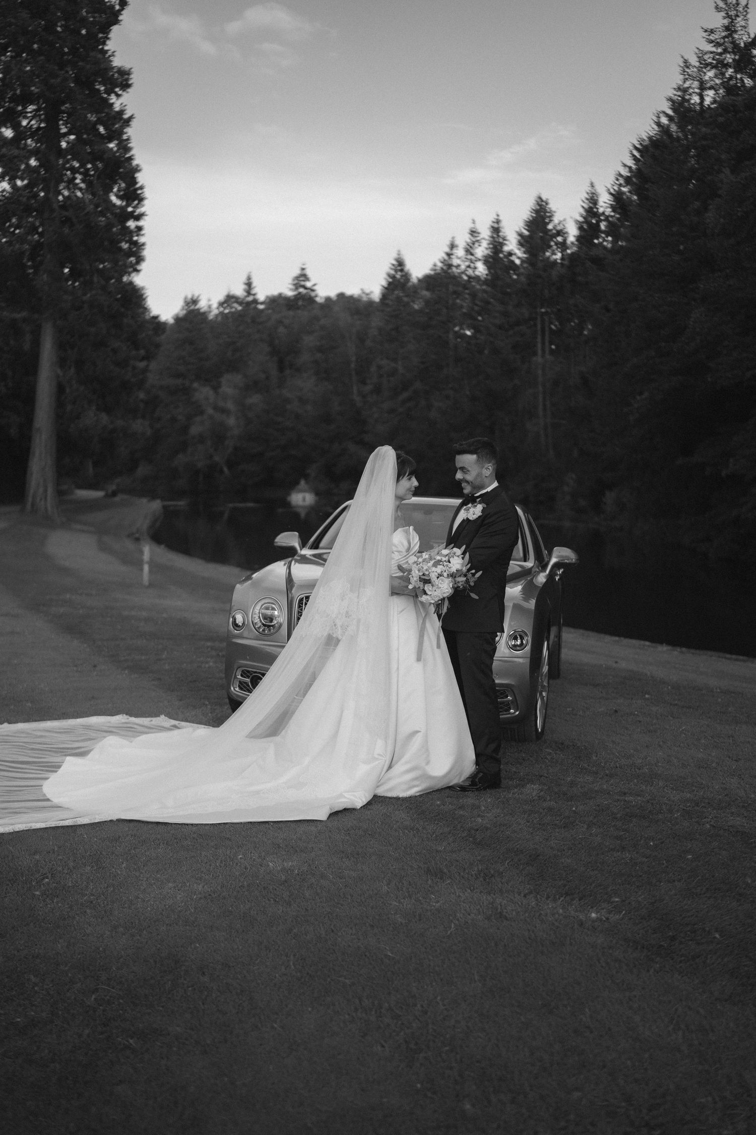 Black and white photo of bride and groom posing by a classic car surrounded by tall evergreen trees.