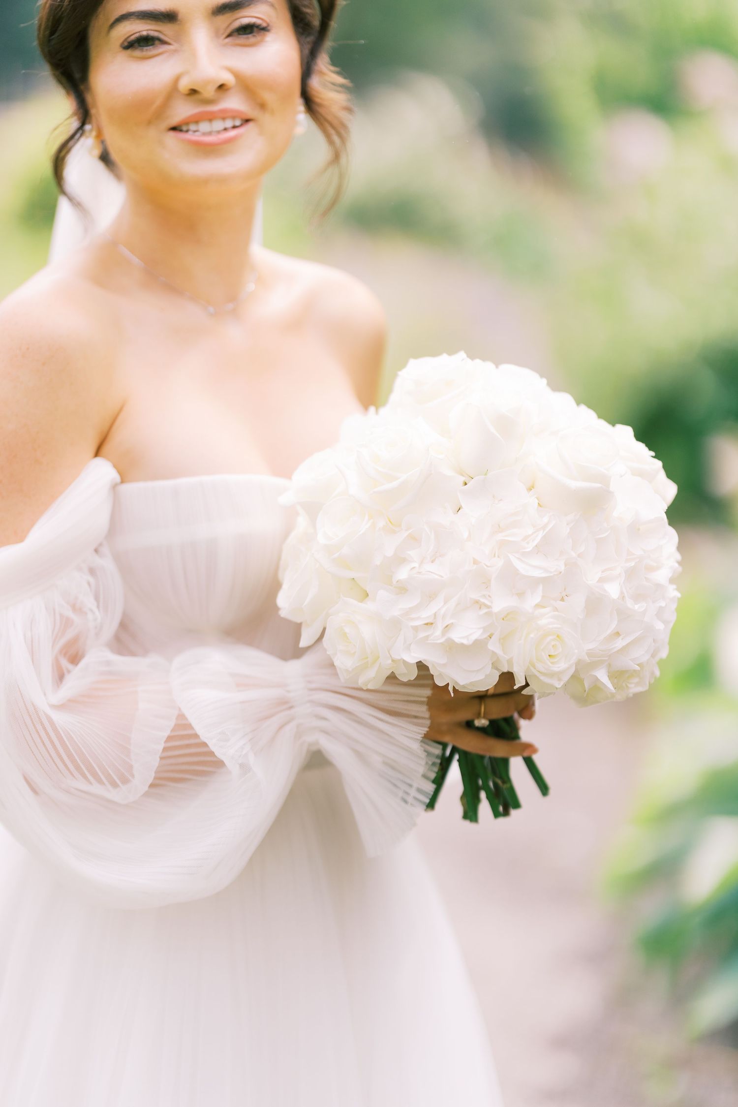Smiling bride in strapless white tulle gown holding elegant white hydrangea and rose bouquet tied with brown ribbon.