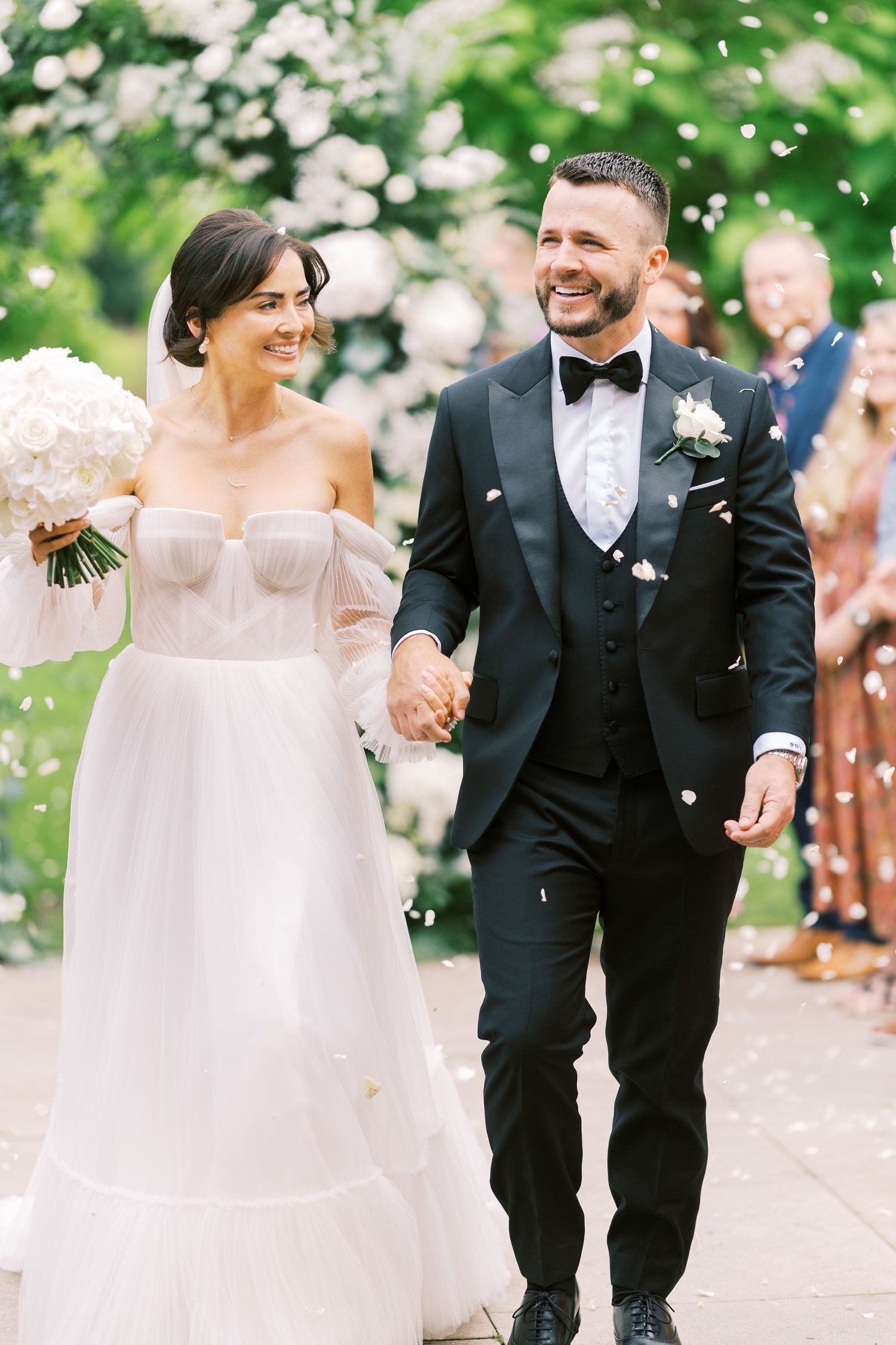 Smiling bride in white off-shoulder gown and groom in black tuxedo walk together as confetti falls at outdoor wedding.