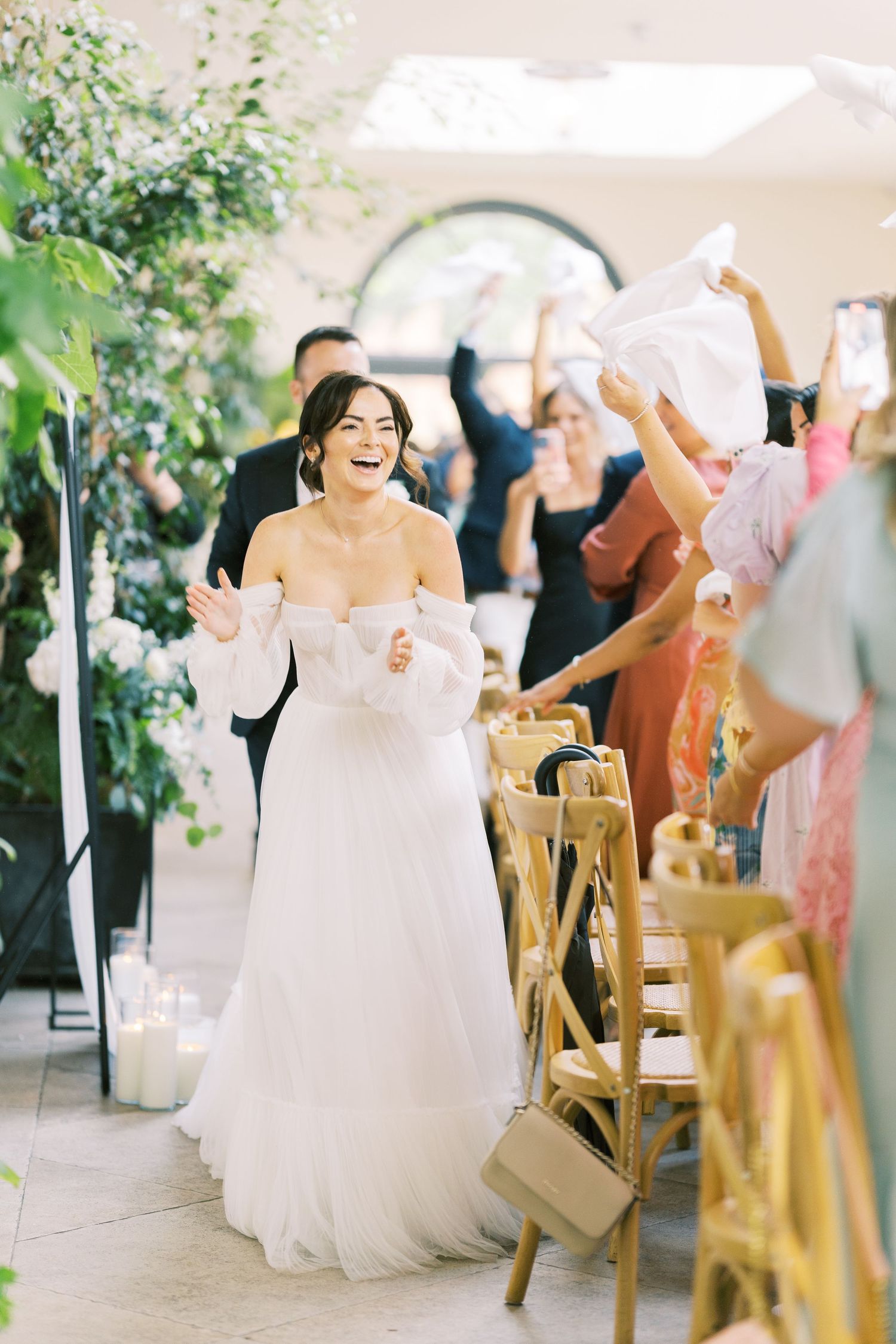 Joyful bride in off-shoulder white gown walks down aisle as guests wave white napkins in elegant venue.