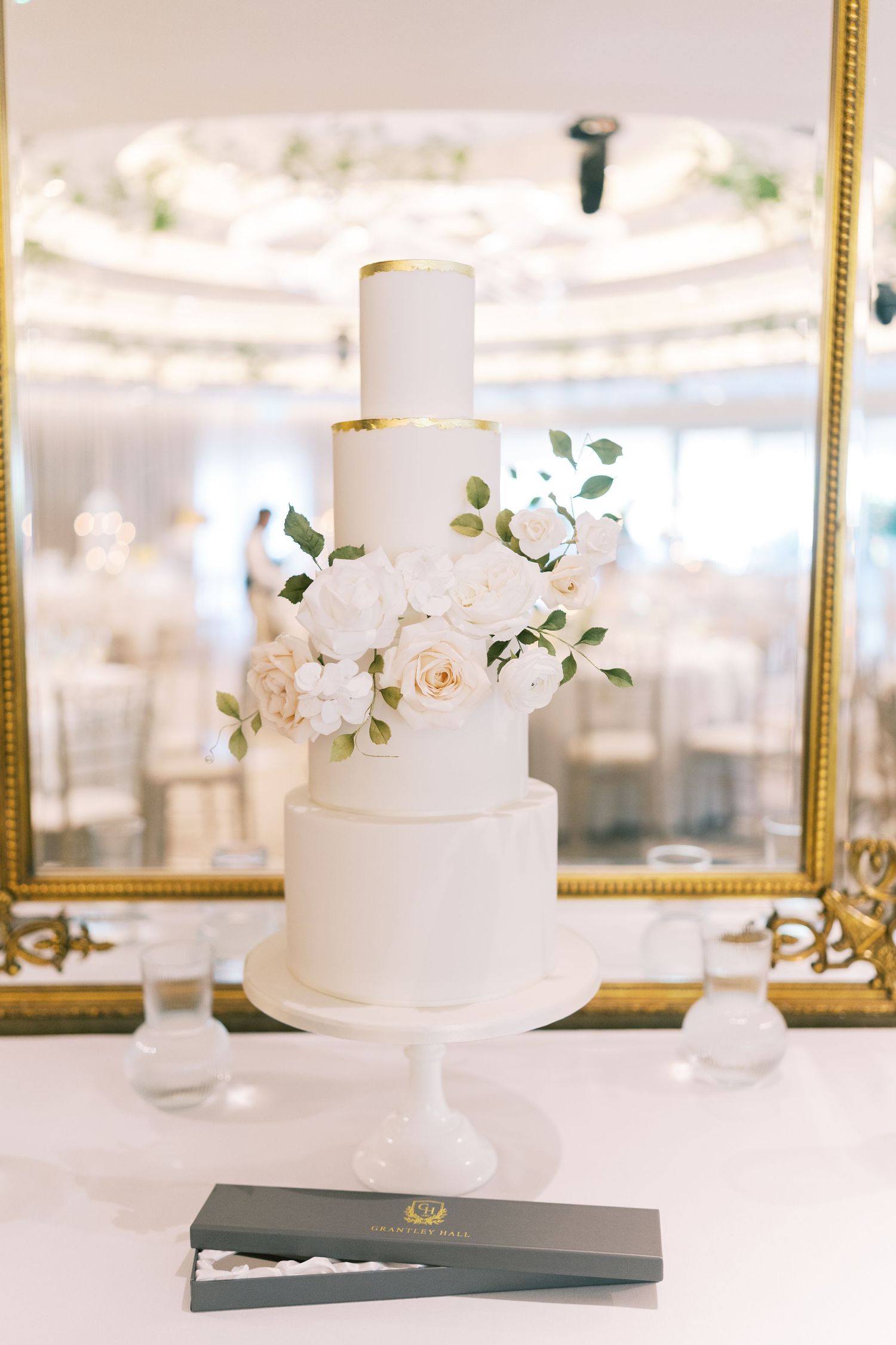 Elegant four-tier white wedding cake with gold accents and fresh white roses on a white cake stand.