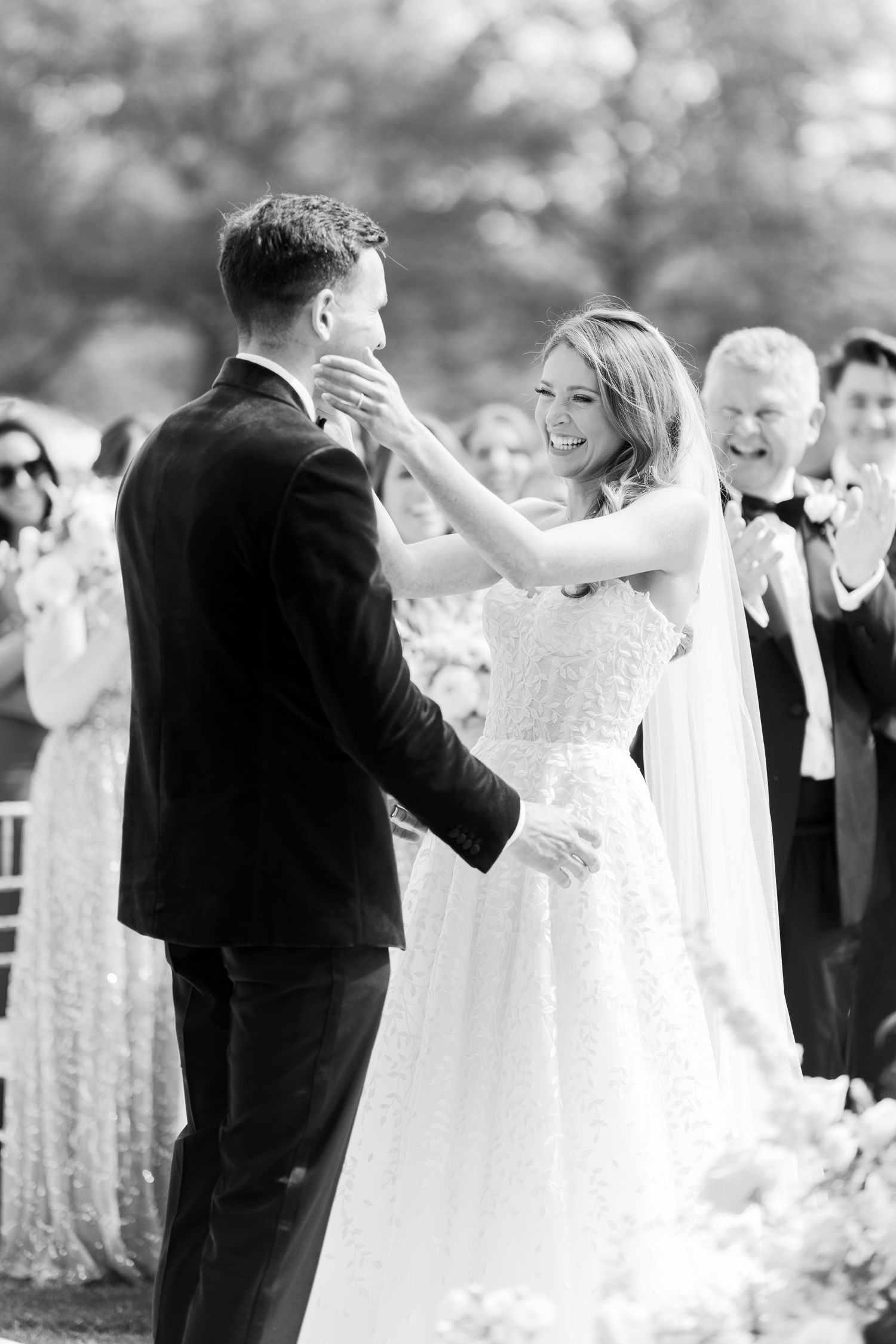 Bride in sparkly gown laughs as she touches grooms face during outdoor wedding ceremony.