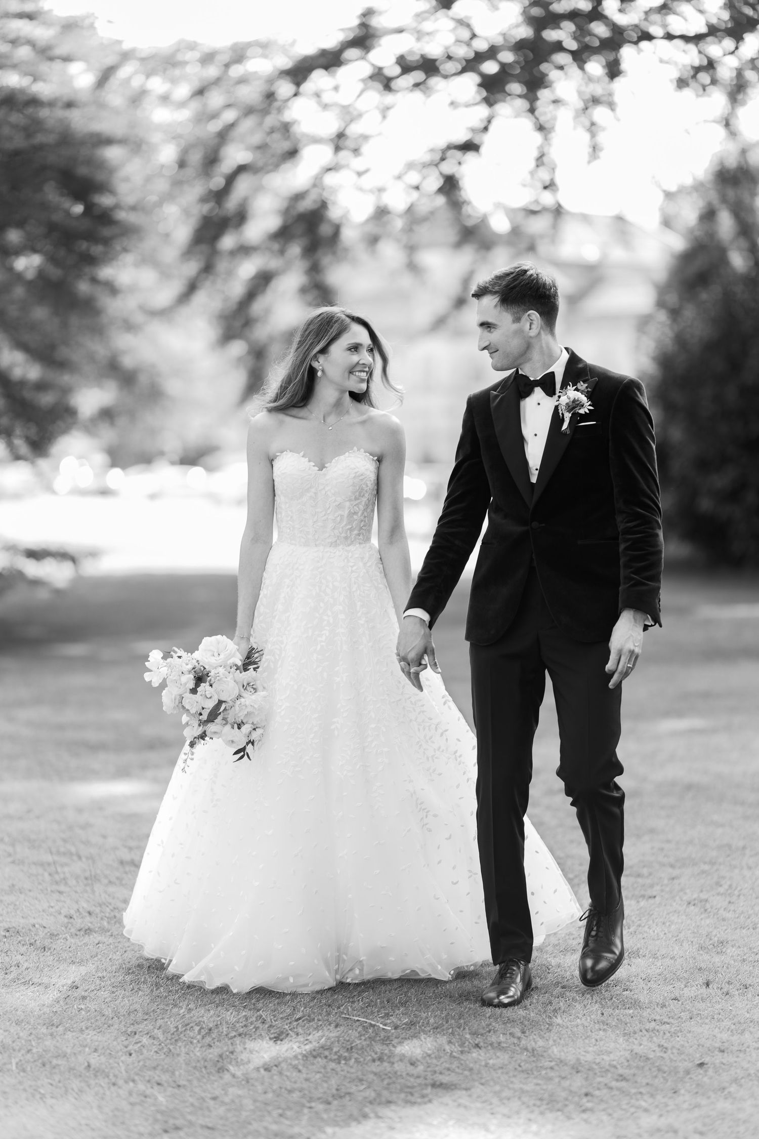 Black and white photo of a smiling bride in a strapless gown and groom in a tuxedo walking hand in hand outdoors.