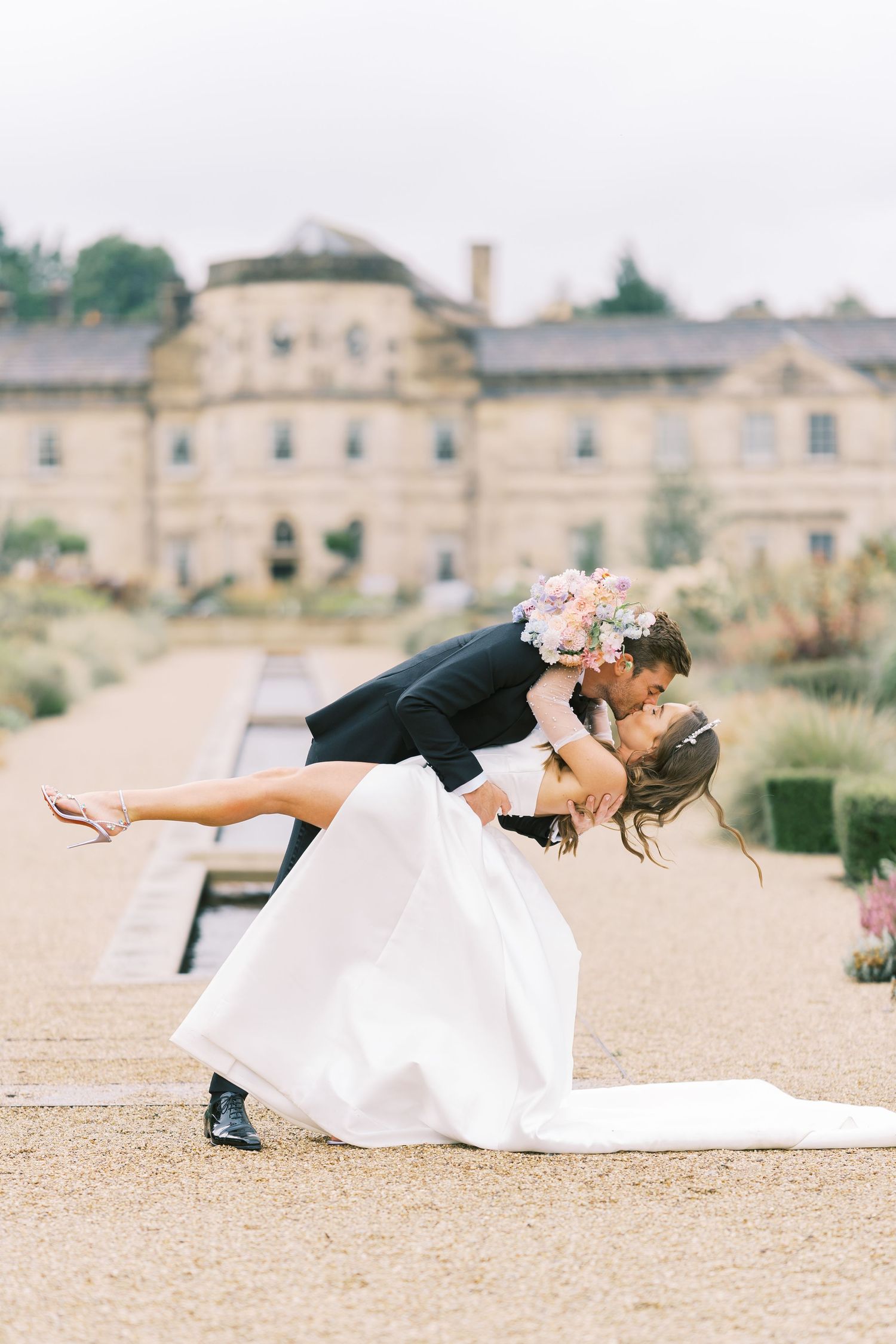 Groom dips bride in a romantic kiss outside a grand stone estate, her pastel bouquet in hand.