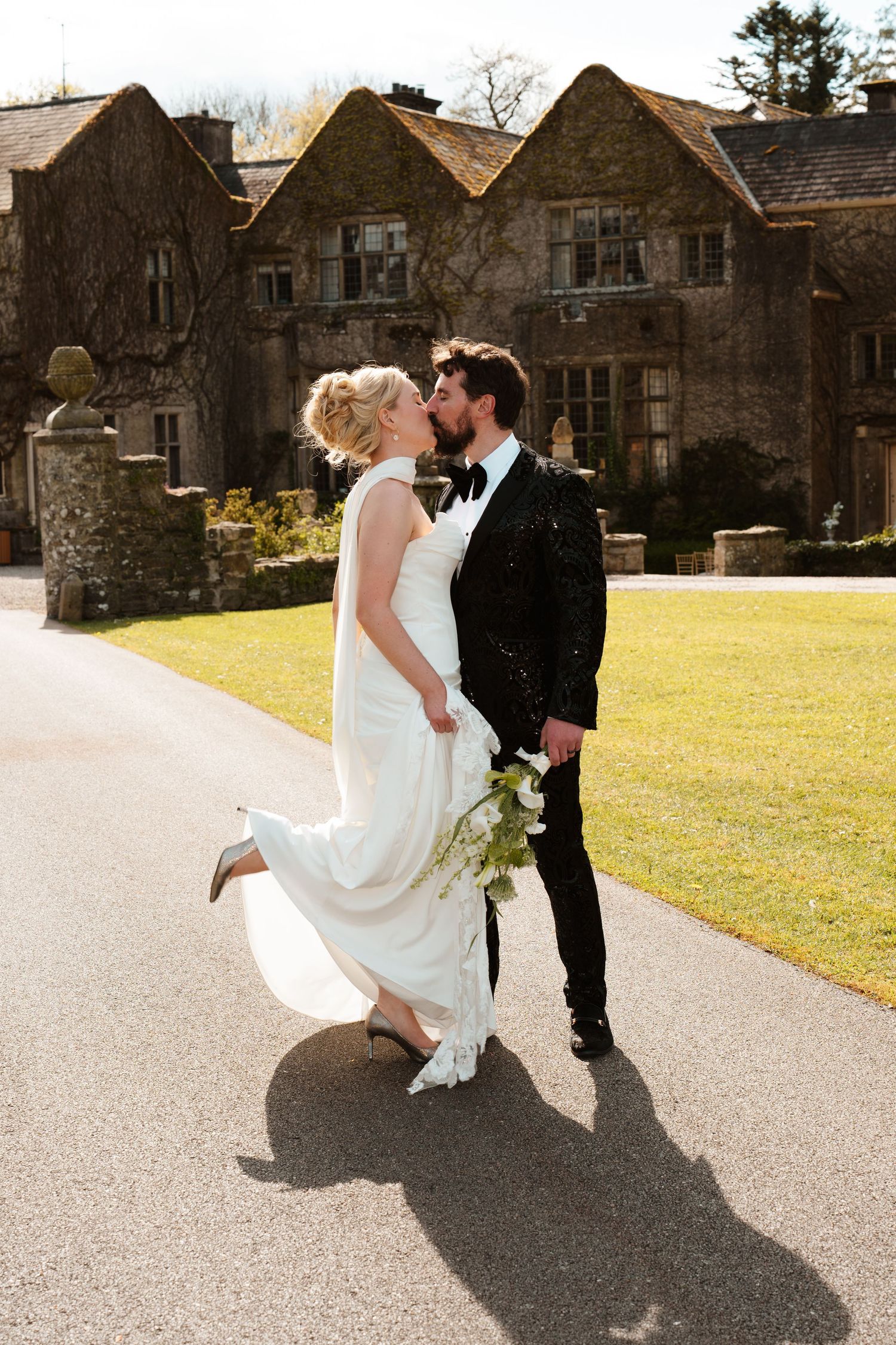 Bride and groom share a romantic kiss while walking outside a stone manor house on their wedding day.