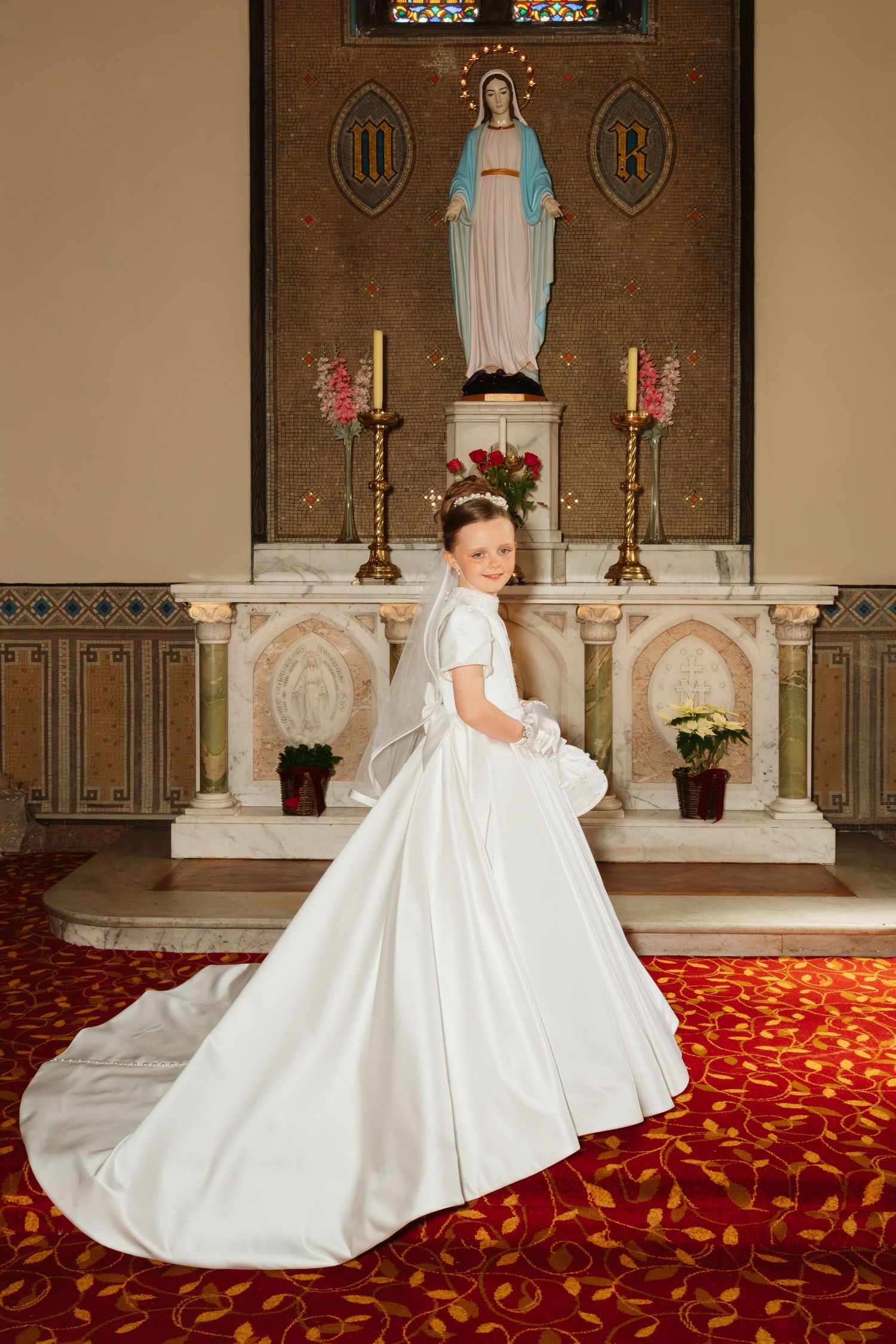 Young girl in white First Communion dress and veil poses before church altar with Virgin Mary statue.