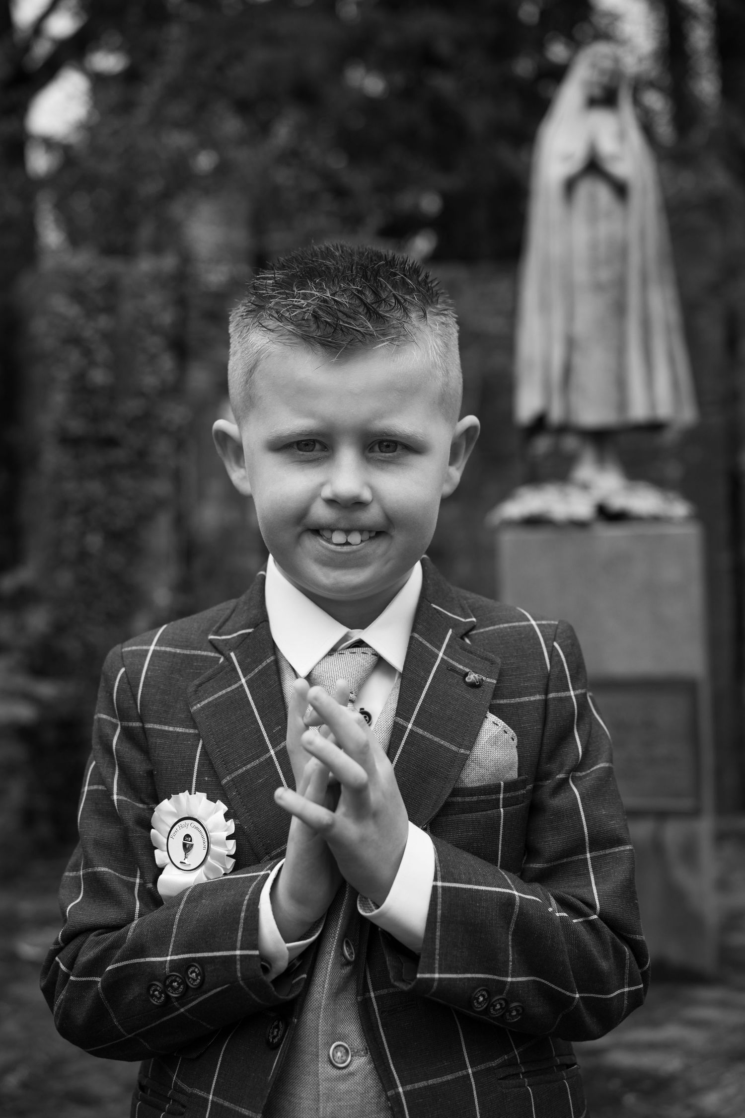 Boy in plaid suit with rosette badge prays hands together near religious statue, black and white First Communion photo.