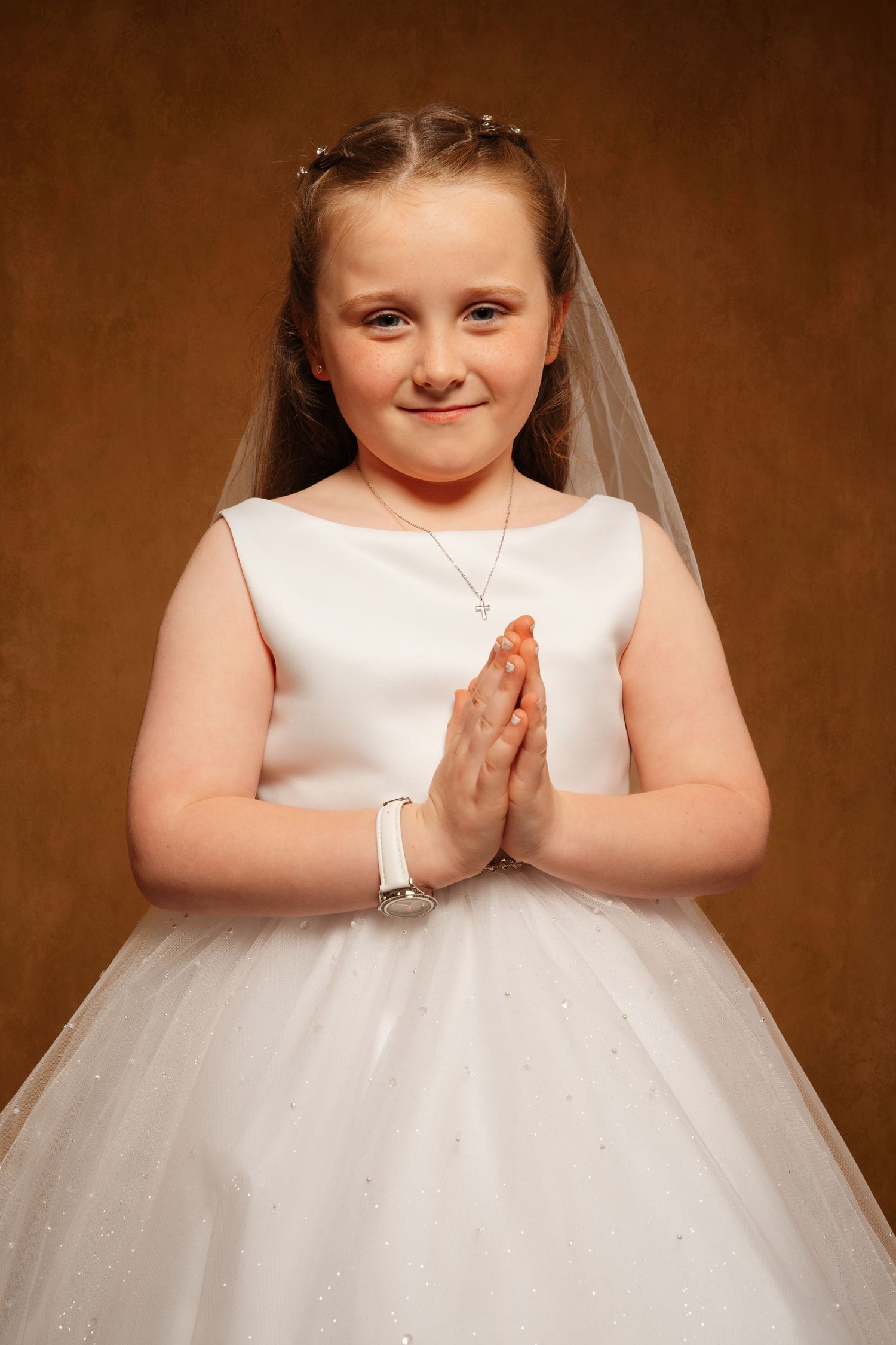 Young girl in white First Communion dress and veil, hands clasped in prayer, smiling against brown background.