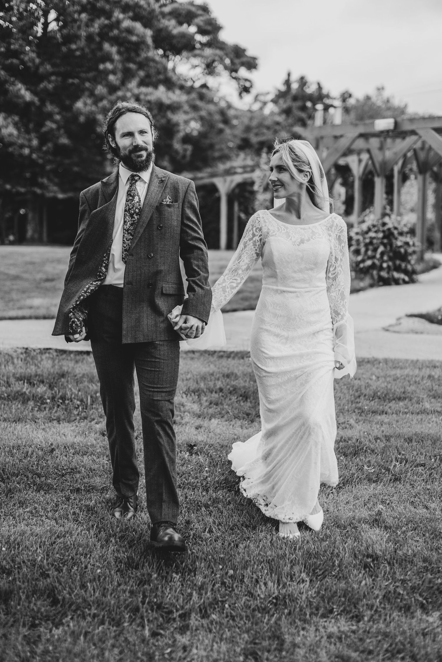 Black and white photo of smiling bride and groom holding hands walking on grass.