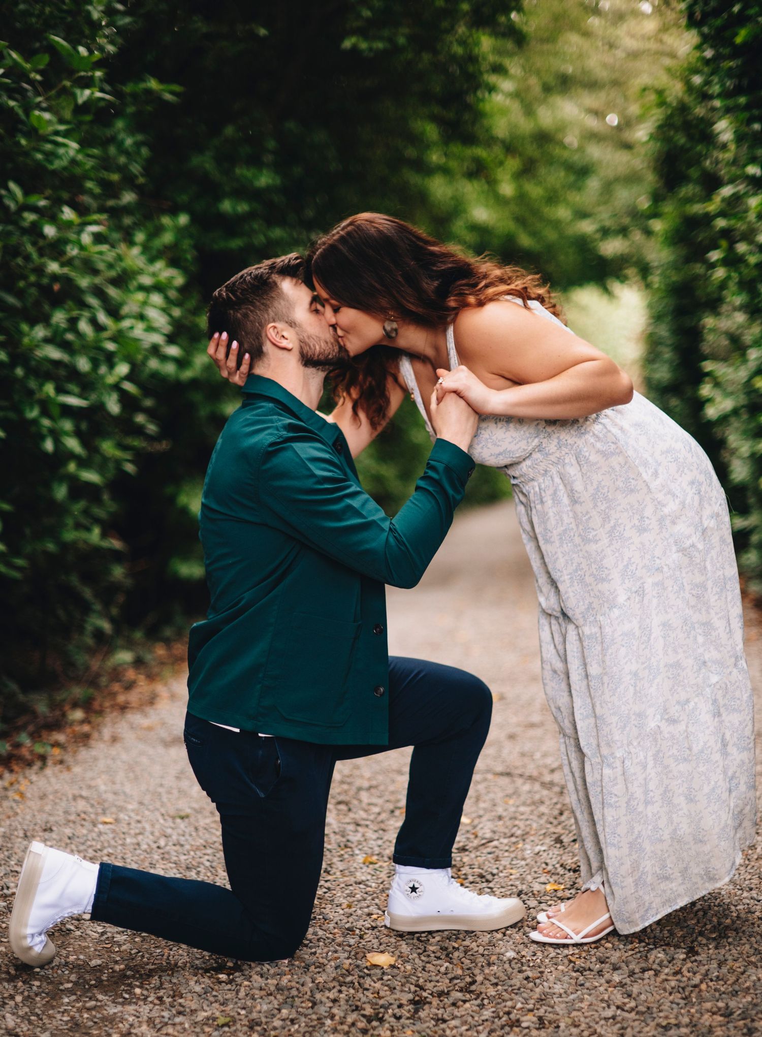 Groom on one knee kissing bride in lace dress along a lush green garden pathway.