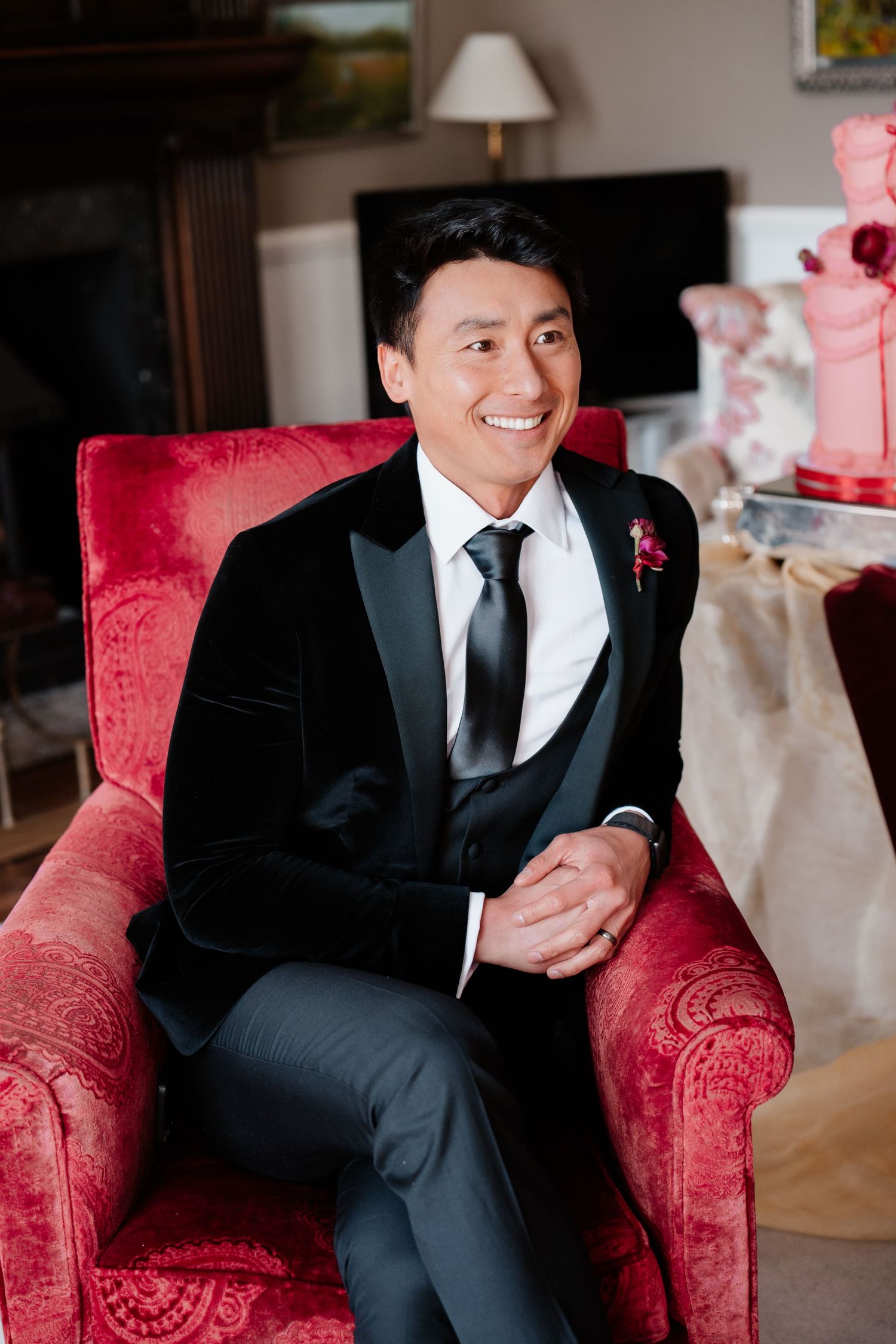Dapper groom in black velvet tuxedo sits smiling in a red velvet chair beside a pink wedding cake.