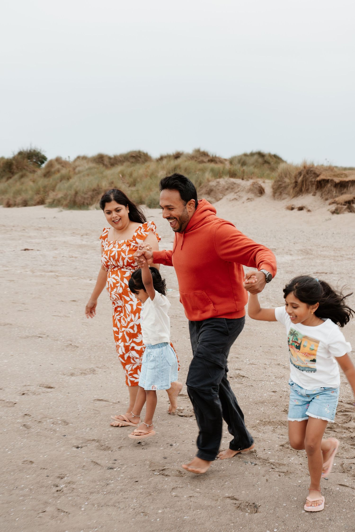 Family of four joyfully playing on a sandy beach, parents holding children's hands, wearing colorful casual clothes.