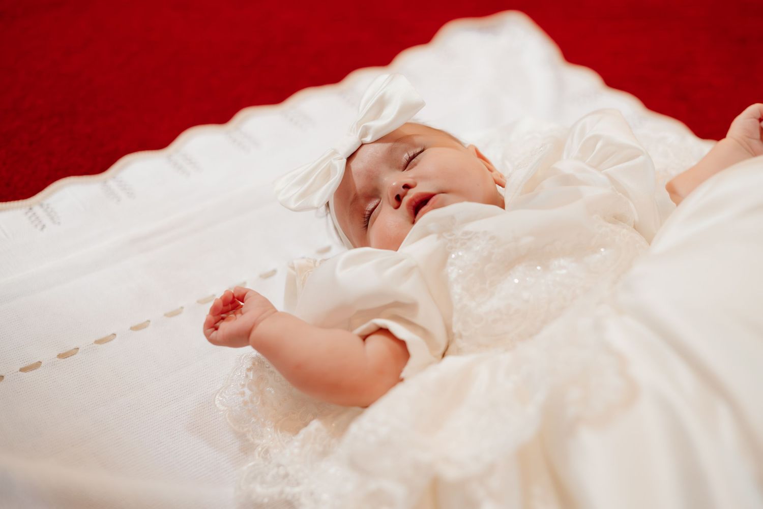 A sleeping newborn baby dressed in a white lace christening gown resting on a white blanket with red background.