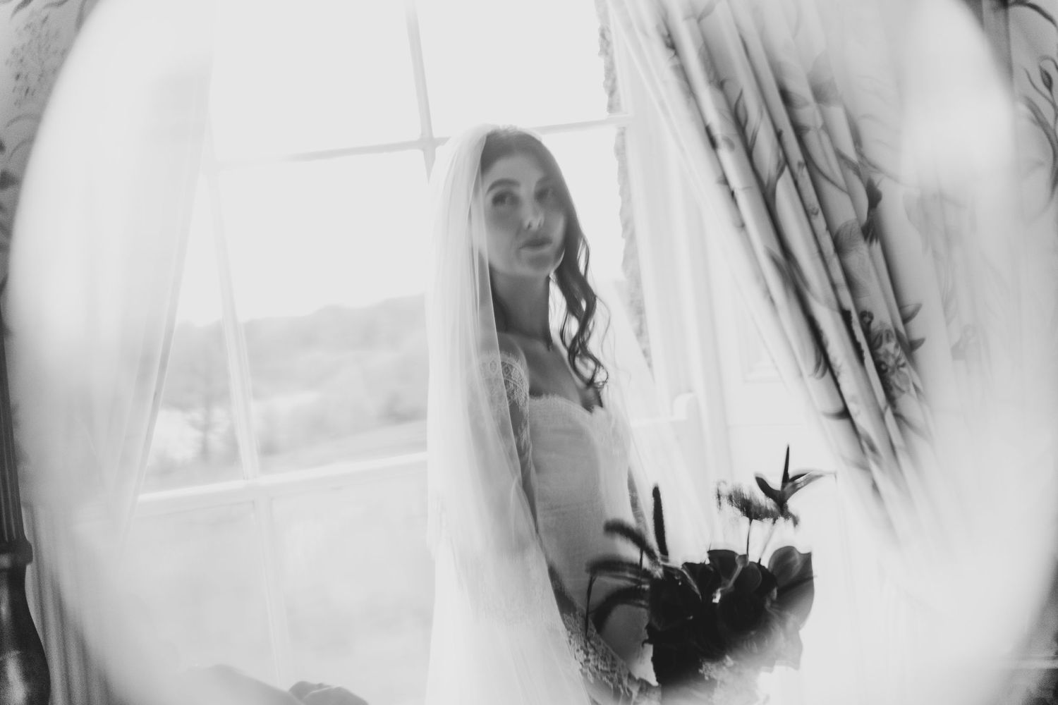 Dreamy black and white bridal portrait through veil with window light and bouquet.