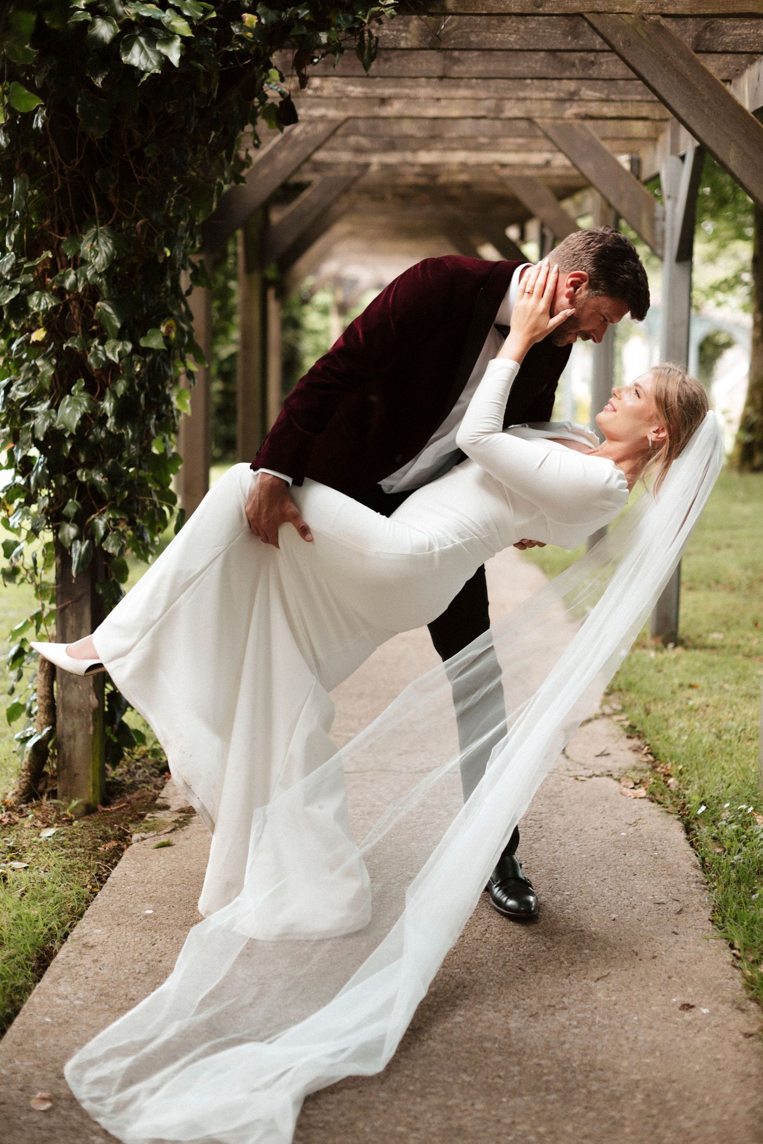 Groom in burgundy velvet suit dips bride in white gown under rustic wooden pergola covered in ivy.