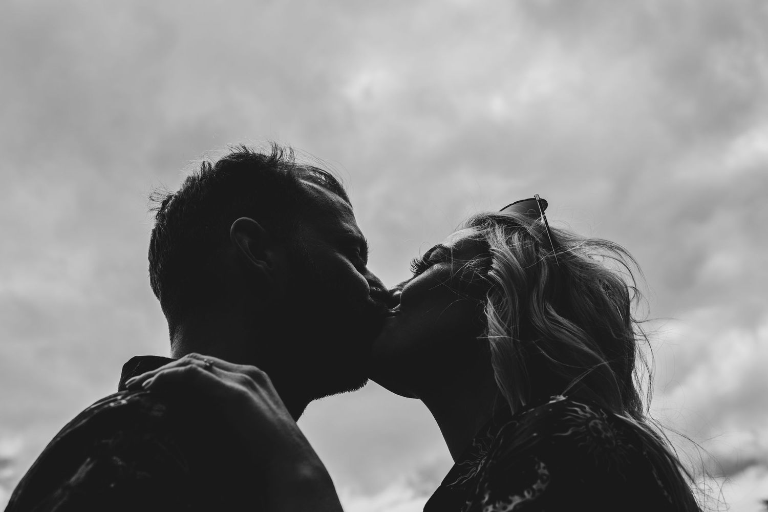 Silhouette of a couple kissing against a dramatic cloudy sky in a romantic black and white photo.