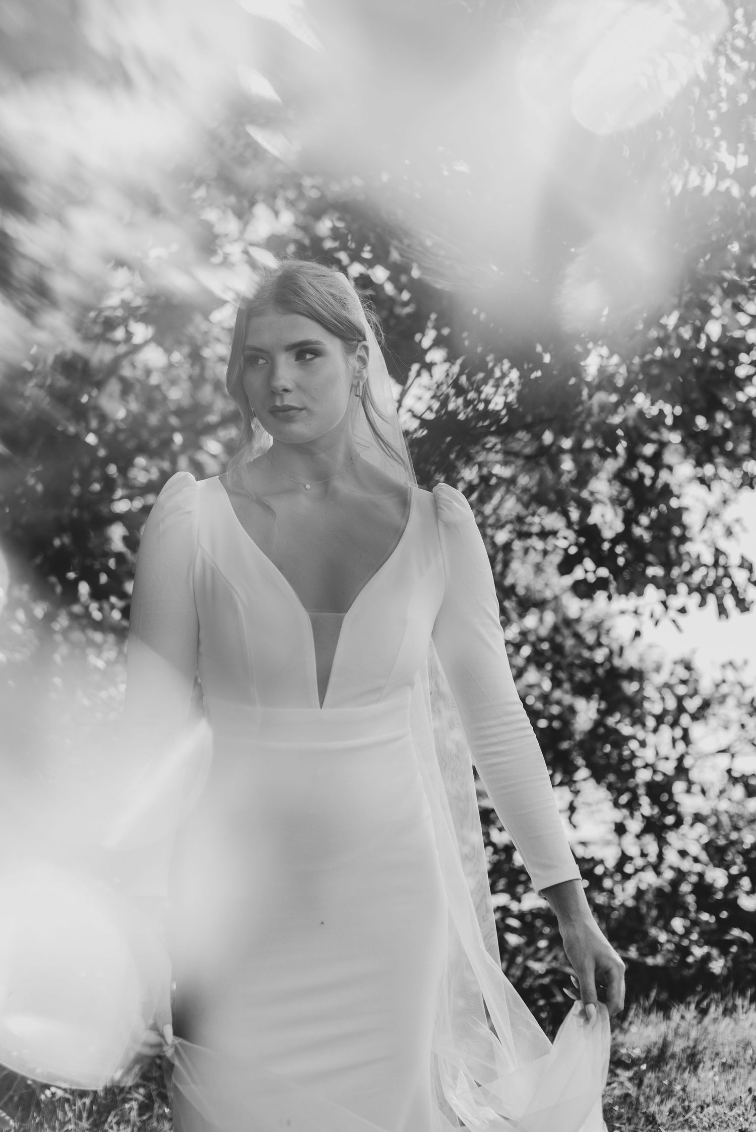 Black and white artistic photo of bride in deep V-neck long-sleeve wedding gown among sunlit trees.