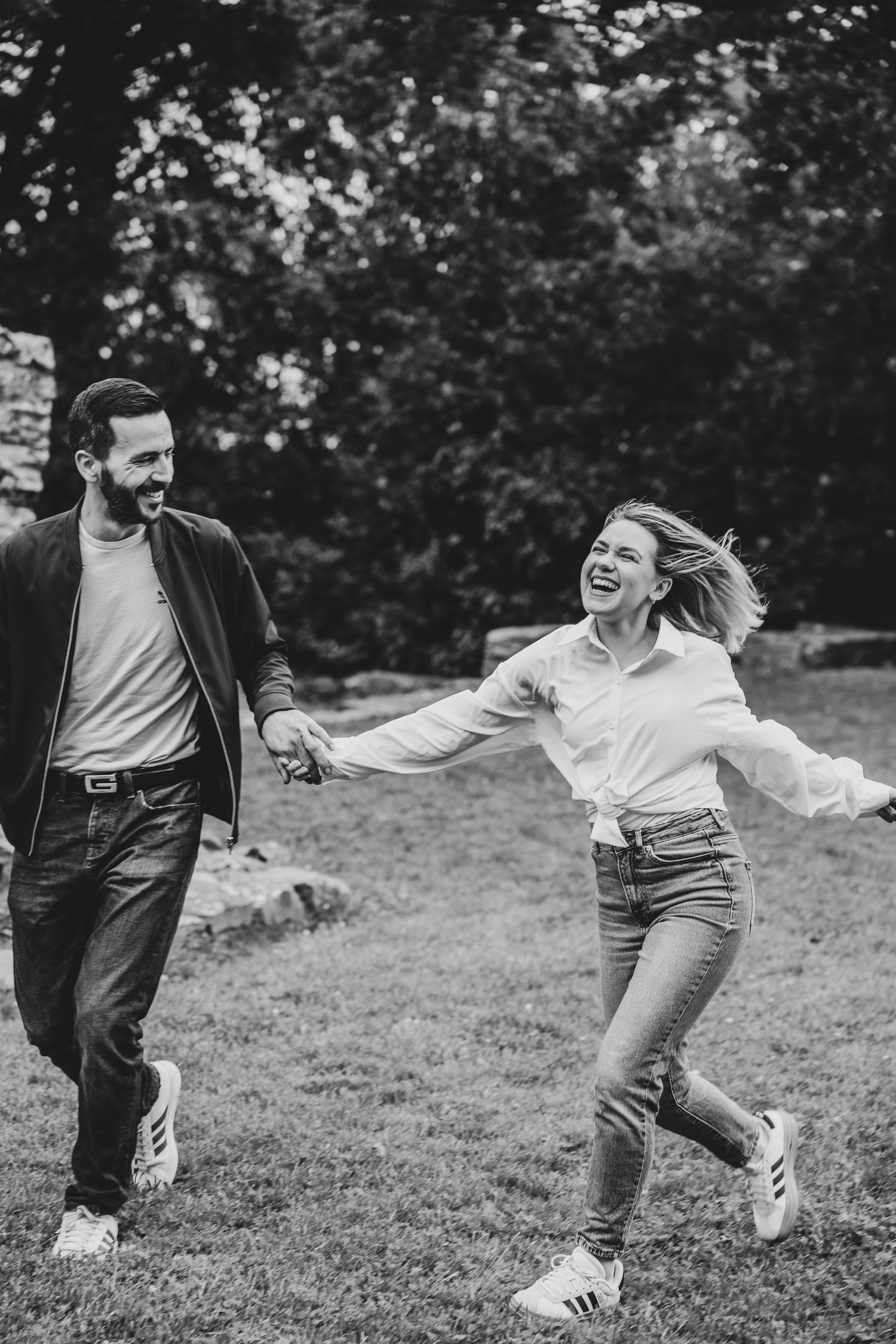 Black and white photo of a joyful couple holding hands and running through a lush green park, laughing together.