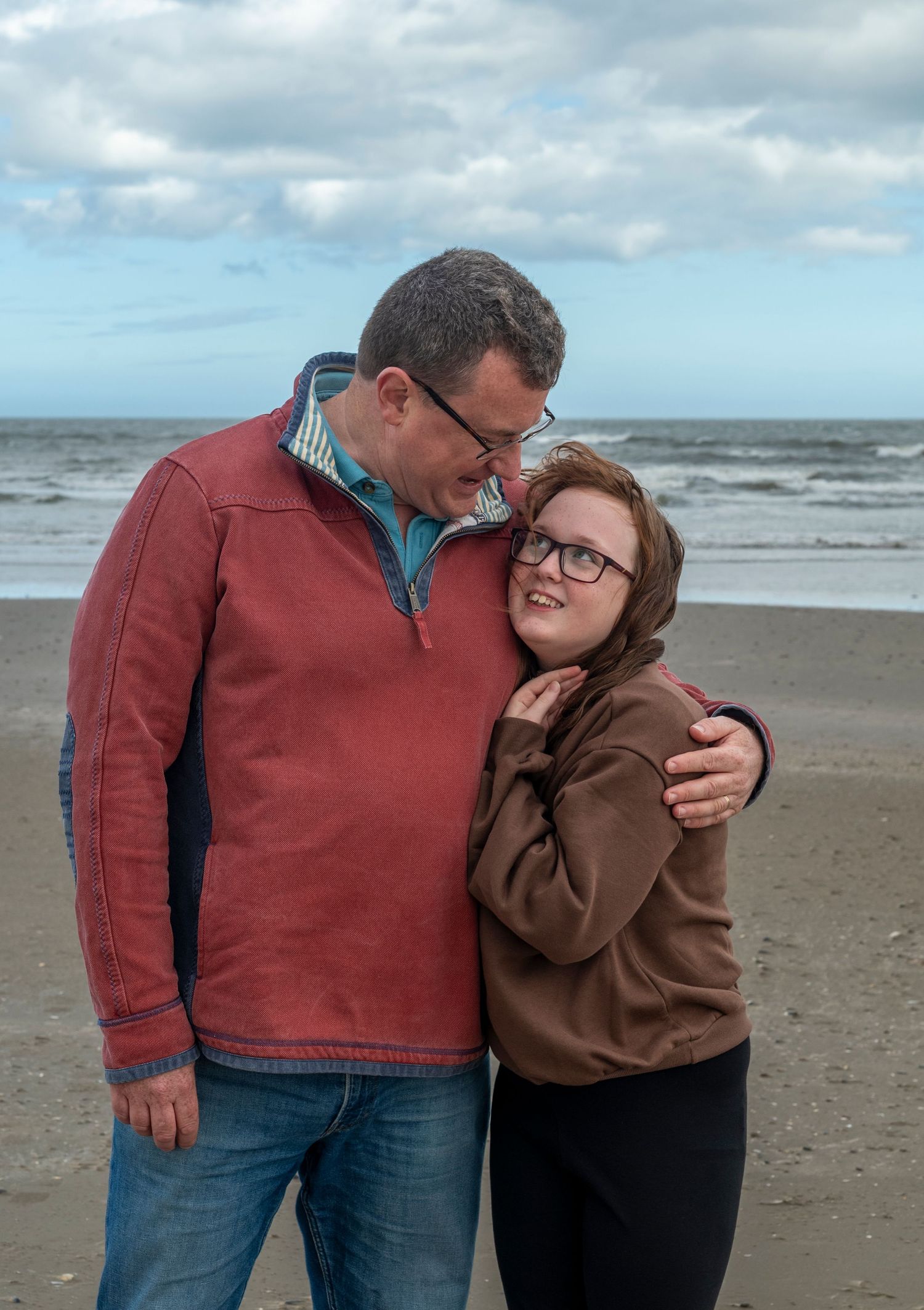 A couple embracing and smiling on a sandy beach with ocean waves and cloudy sky behind them.