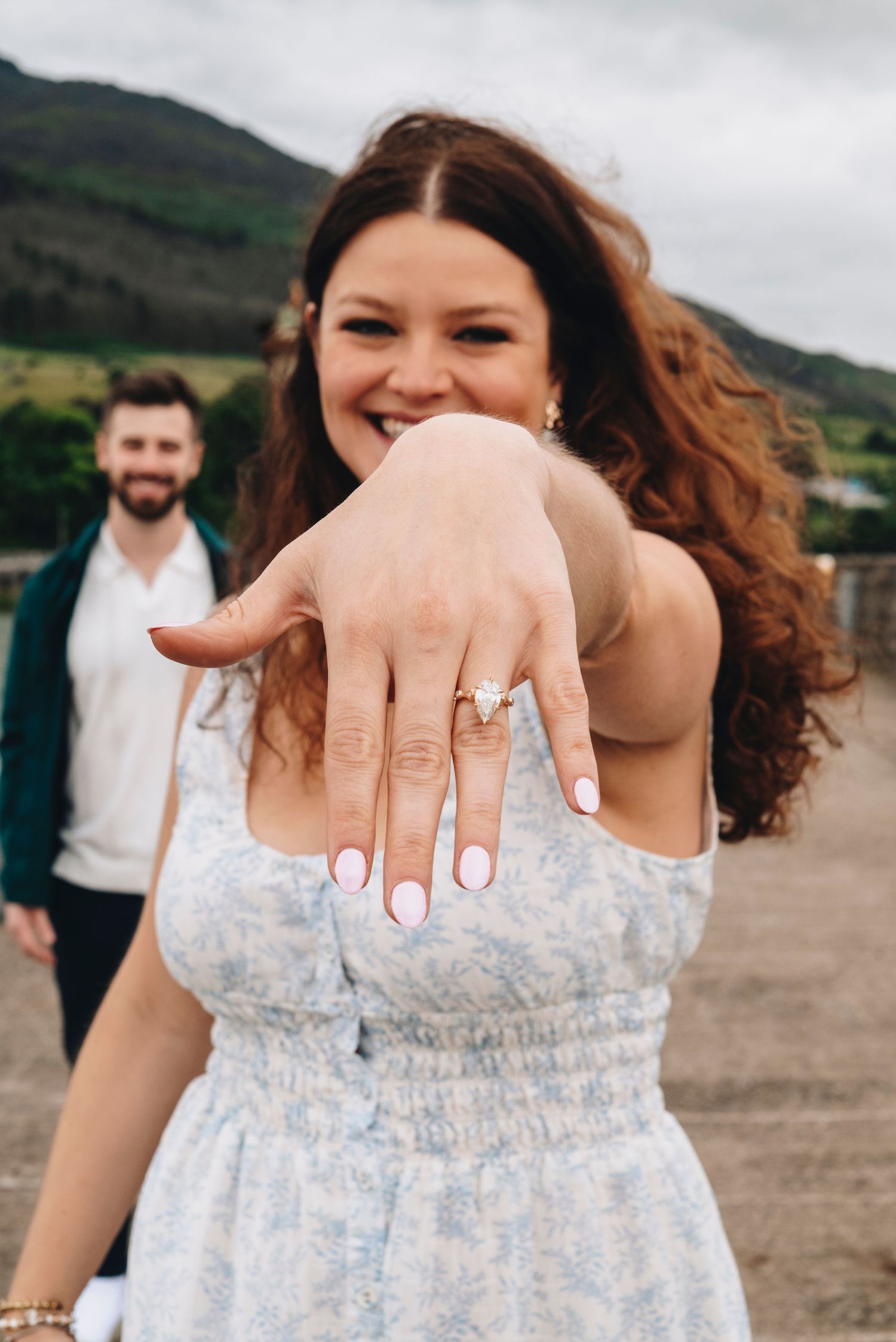 Smiling bride showing off her engagement ring with groom standing behind her.