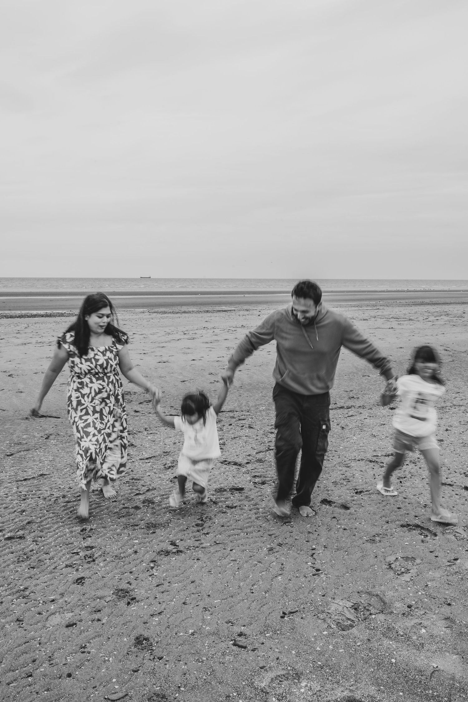 Black and white photo of a happy family of four walking together on a beach, holding hands near the ocean.