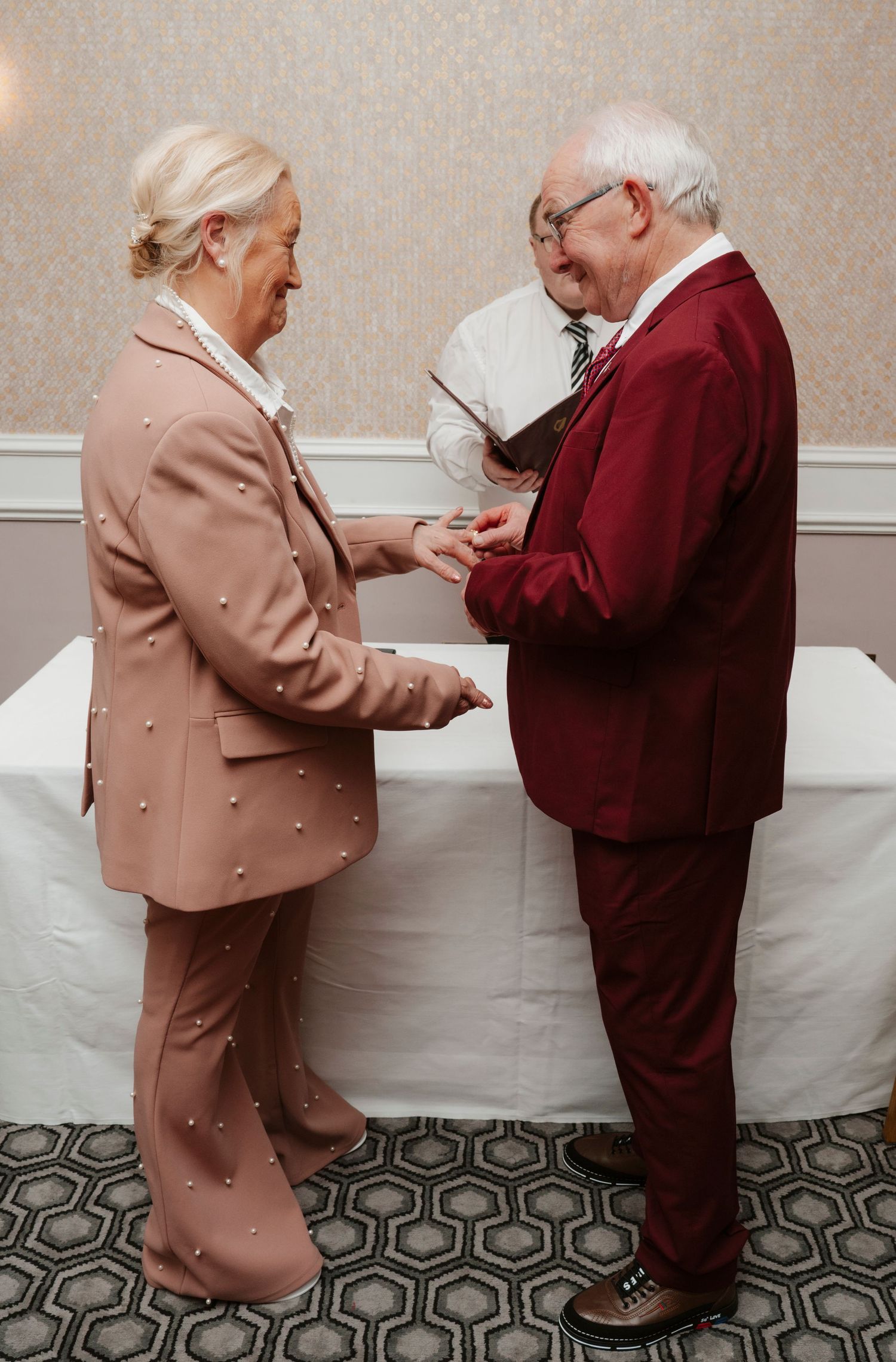 Elderly couple exchanging rings during wedding ceremony, woman in pink pearl suit, man in burgundy suit.