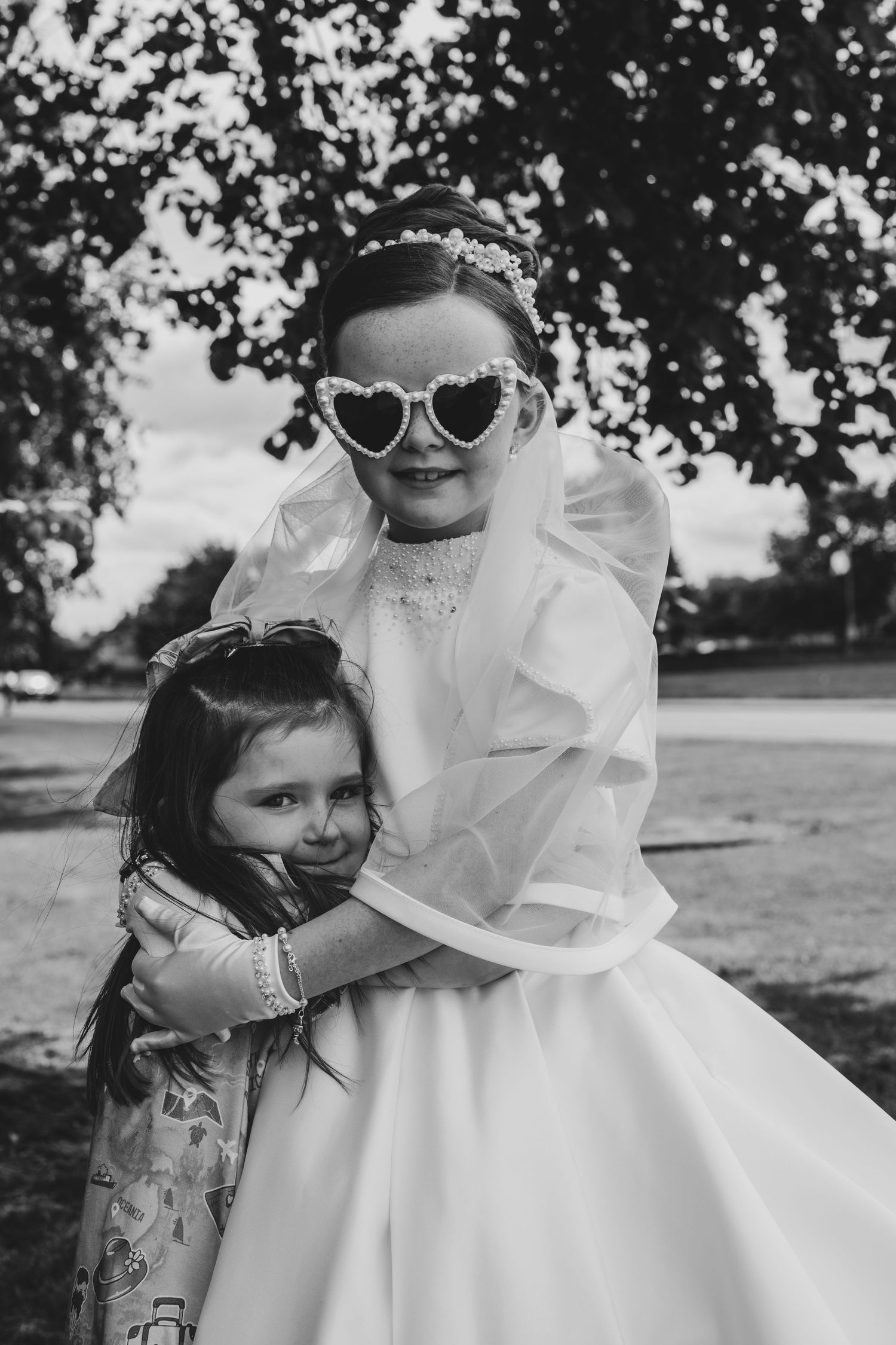 Black and white photo of two young girls hugging outdoors, one wearing a white dress, veil, and heart-shaped sunglasses.