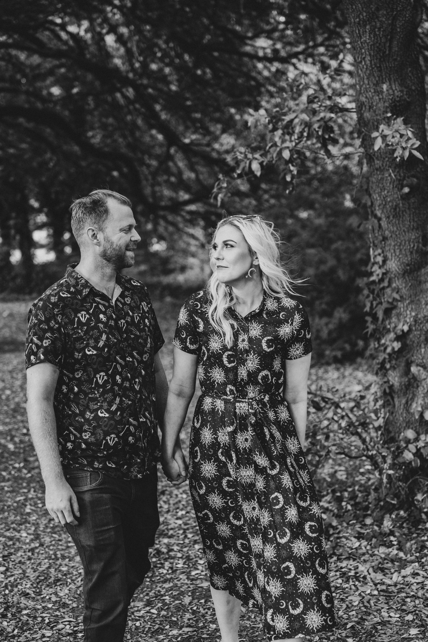 Black and white photo of a couple holding hands and smiling at each other among autumn trees.