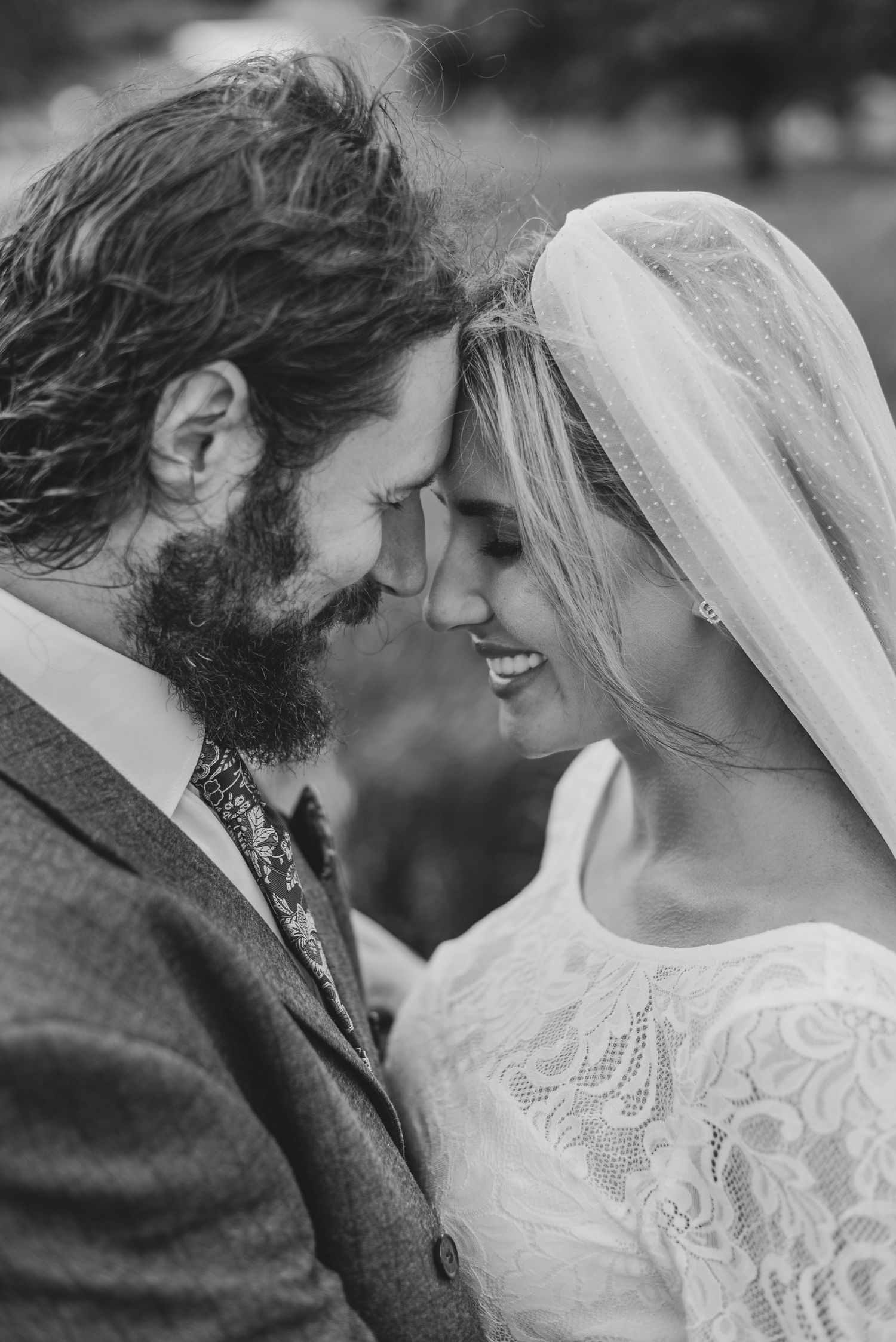 Black and white wedding photo of bearded groom and smiling bride in lace dress and veil touching foreheads romantically.