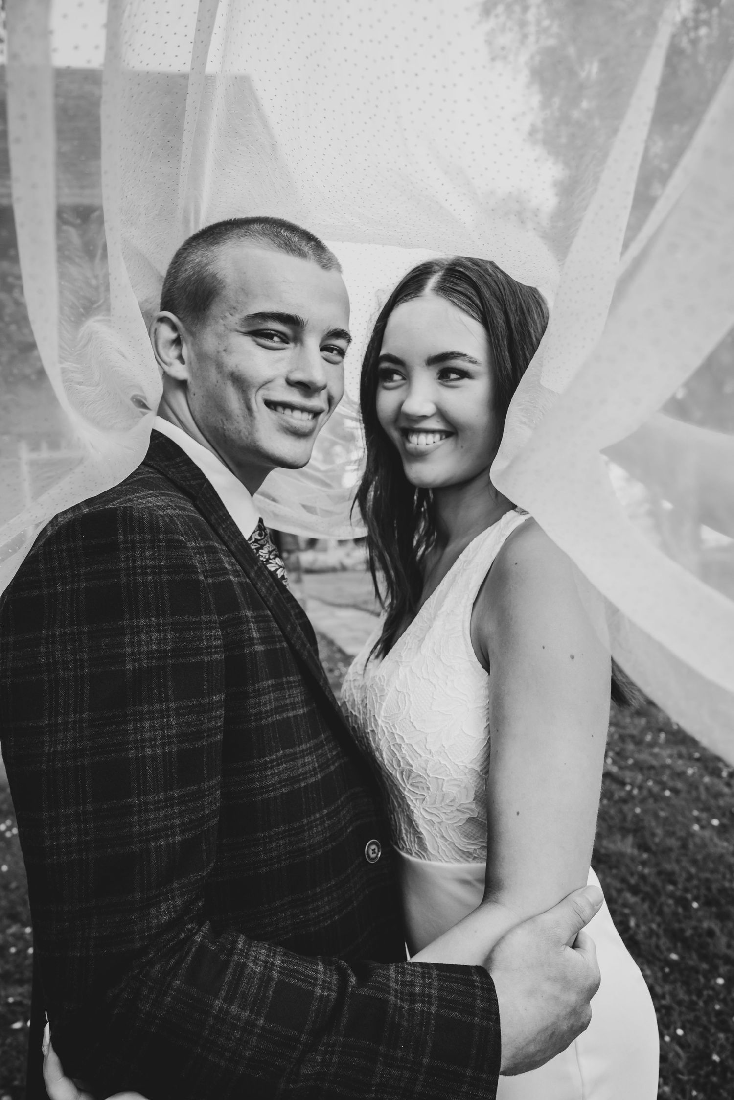 Black and white photo of smiling bride and groom embracing under flowing veil.
